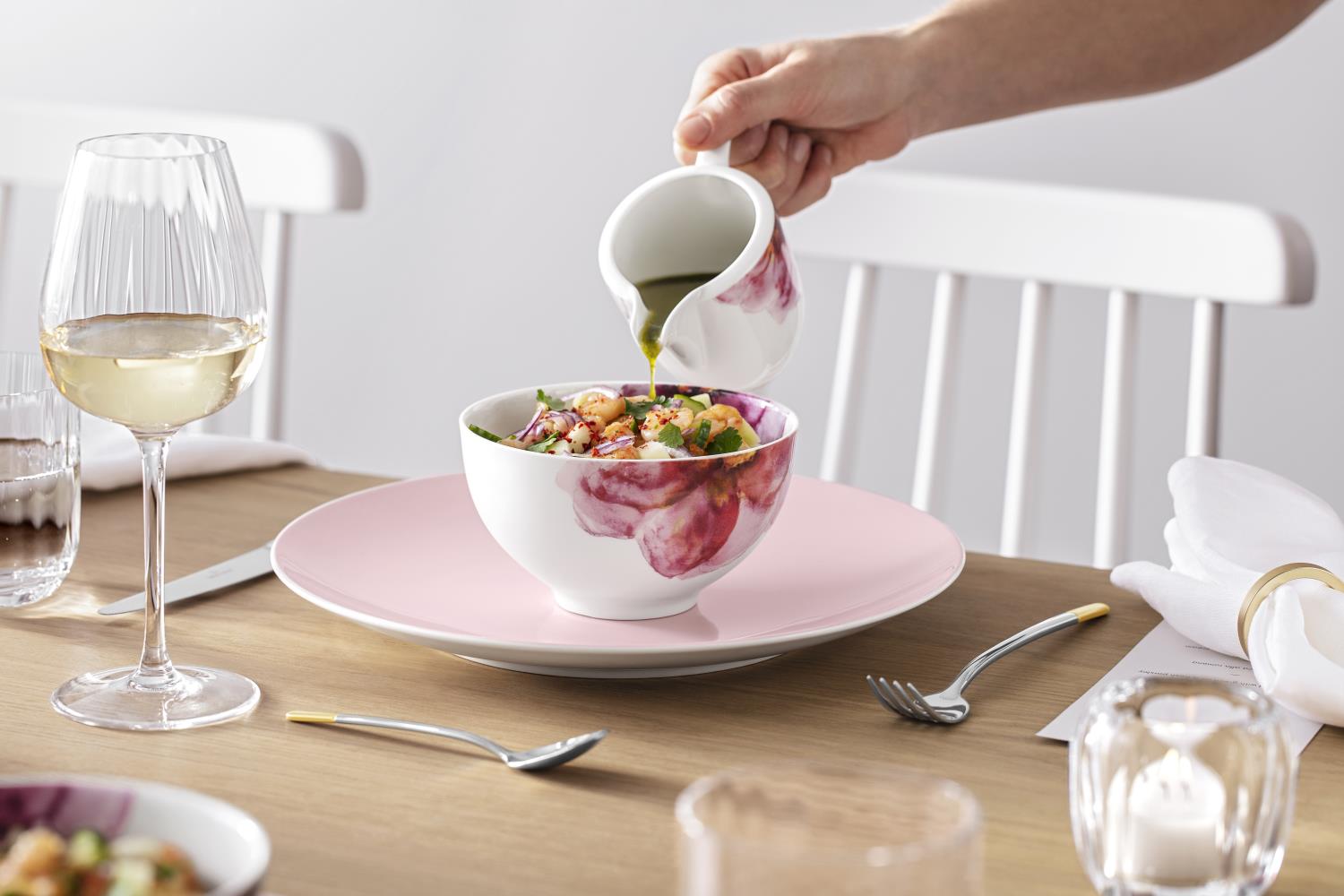 Eine Hand gießt Öl aus einer Kanne in eine Schüssel mit Salat, daneben ein Glas Weißwein und ein Tel