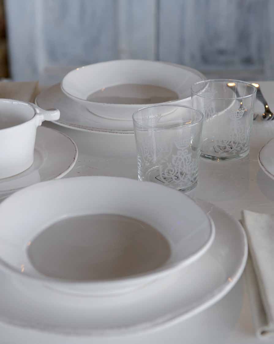 Tischgedeck mit weißen Tellern, Gläsern und Besteck auf einem hellen Tisch. Im Hintergrund sind unscharfe Wände zu sehen.