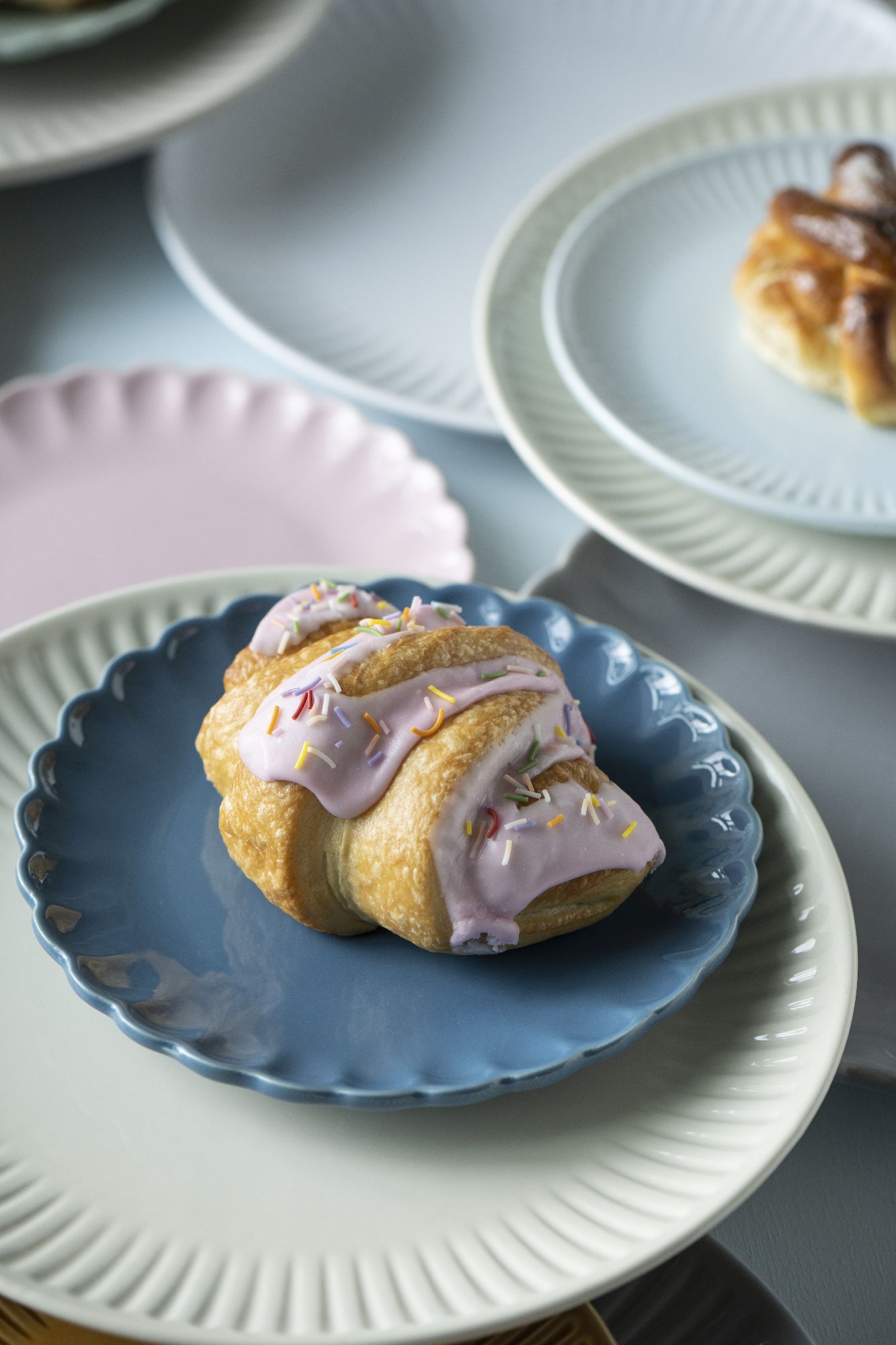Ein Croissant mit rosa Zuckerguss und bunten Streuseln auf einem blauen Teller, umgeben von weiteren Tellern.