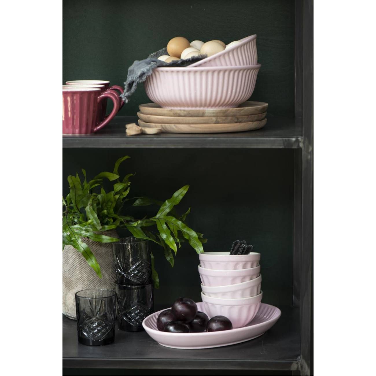 Regal mit Geschirr: rosa Schalen, Tassen, Gläser, Holzuntersetzer und eine Pflanze. Eier und Pflaumen sind sichtbar.