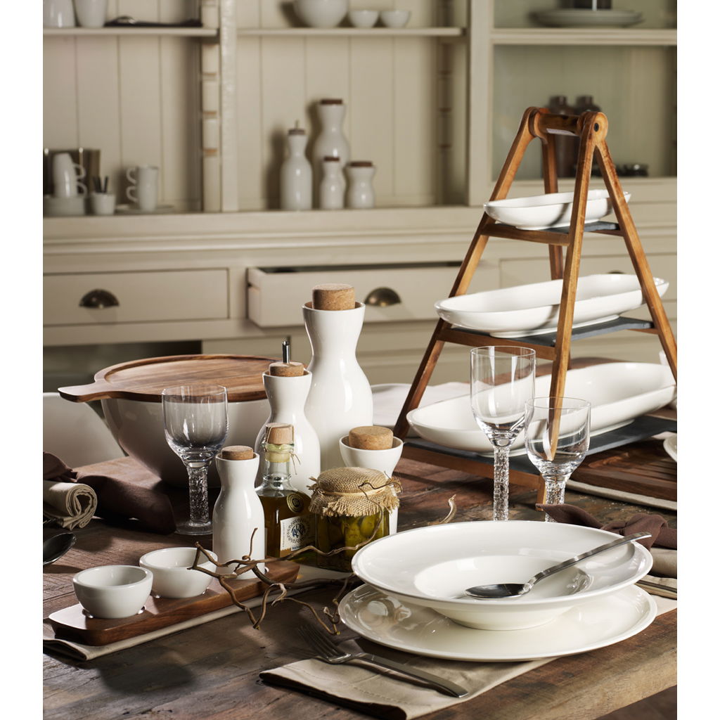 Tischgedeck mit weißen Tellern, Gläsern, Karaffen und einer Holzschale auf einem Holztisch. Im Hintergrund Geschirr im Regal.