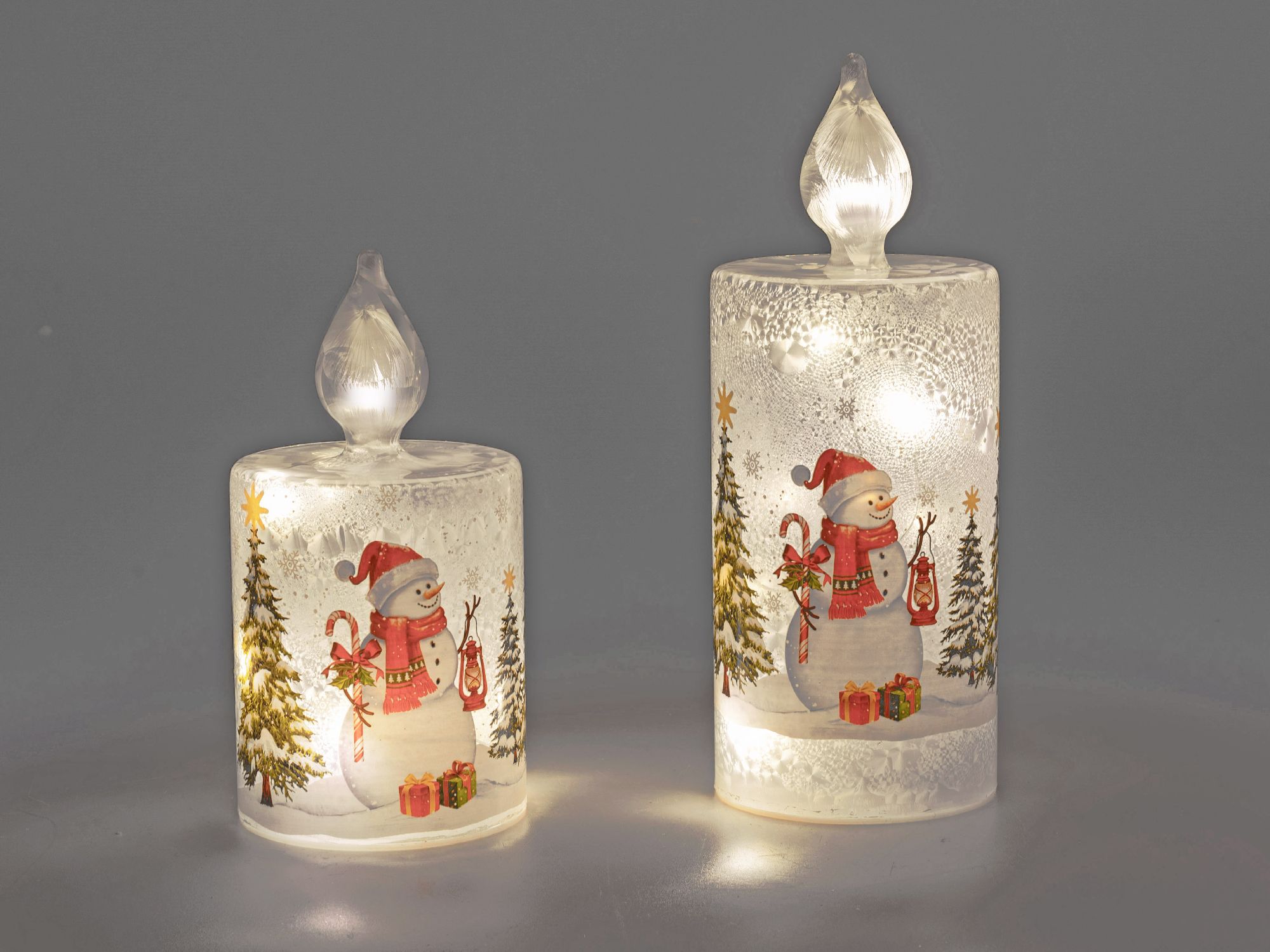Zwei leuchtende Kerzen mit Schneemann-Motiven und Weihnachtsbäumen, auf grauem Hintergrund.