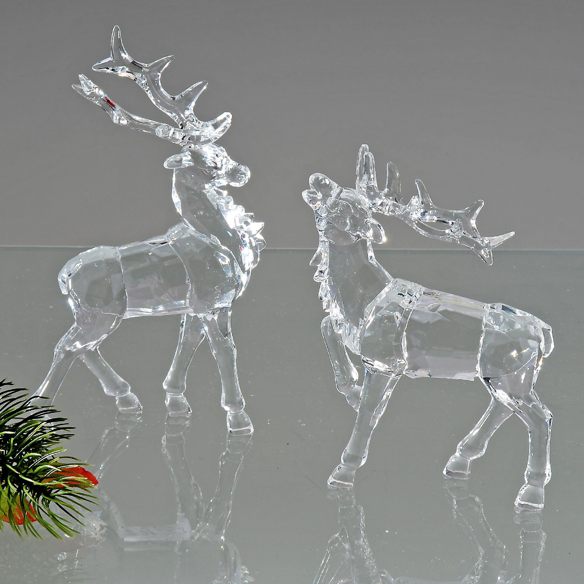Zwei durchsichtige, gläserne Rehe stehen auf einer reflektierenden Oberfläche, umgeben von einem grü