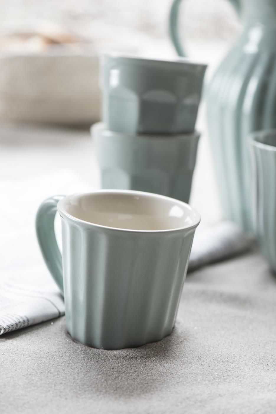 Eine hellblaue Tasse steht auf einem Tisch, umgeben von weiteren Tassen und einem Kännchen. Der Tisch ist mit einem Tuch bedeckt.