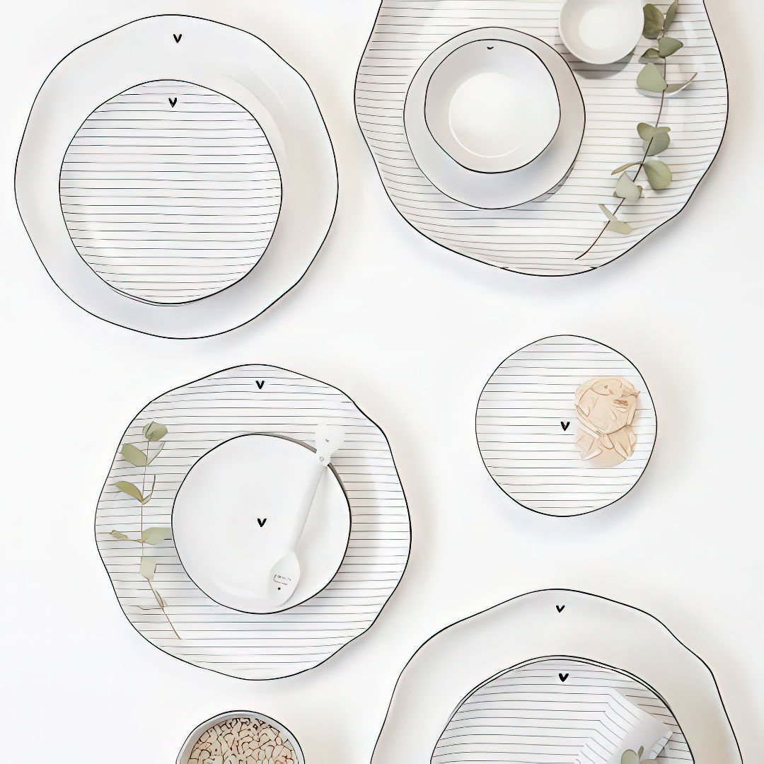 Mehrere Teller und Schalen mit gestreiftem Muster und einem kleinen Zweig liegen auf einem weißen Untergrund.