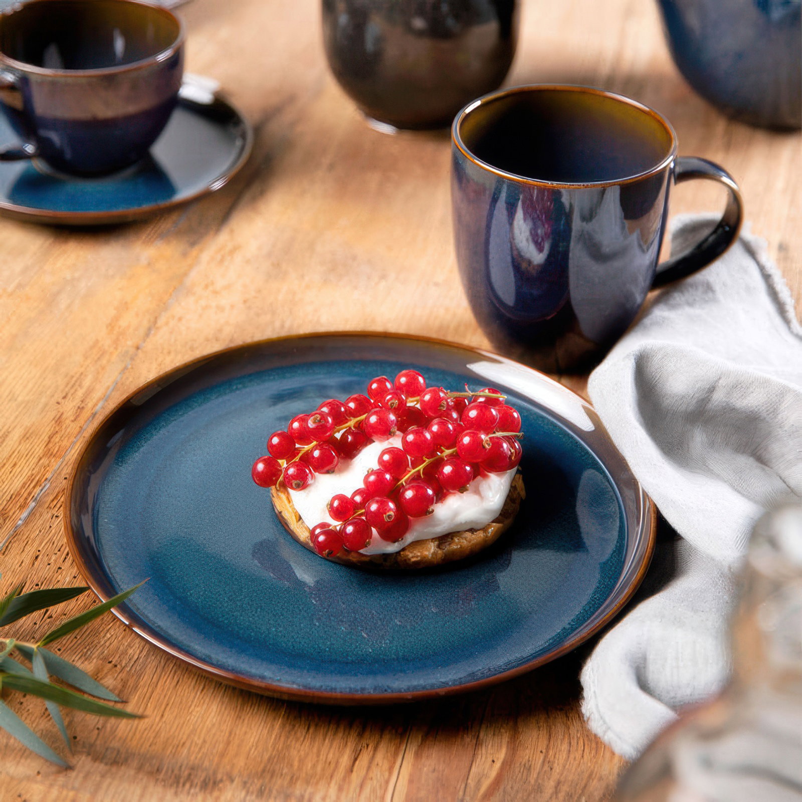 Ein blauer Teller mit einem Keks, belegt mit Sahne und roten Johannisbeeren, steht auf einem Holztisch. Tassen und ein Tuch sind sichtbar.