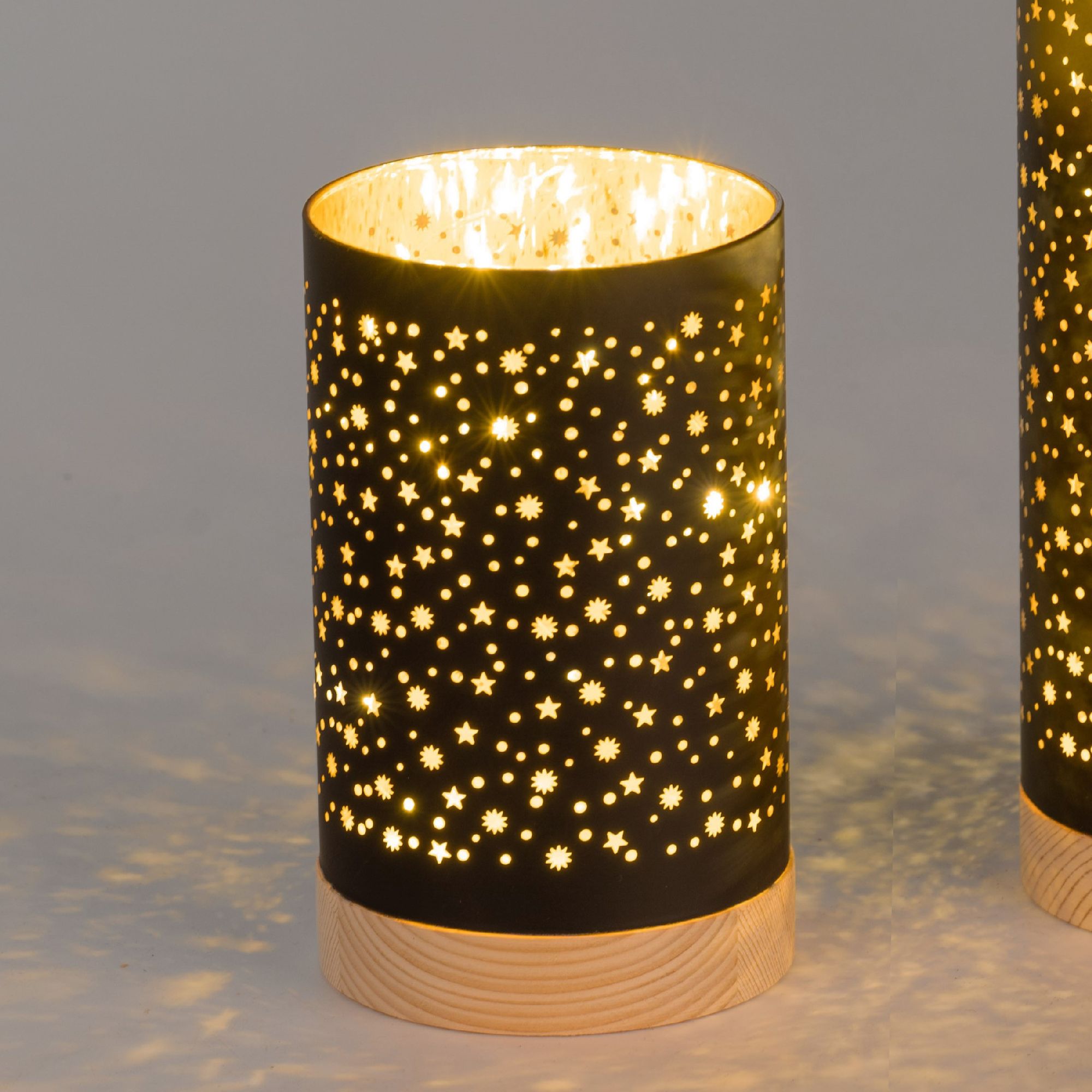 Schwarze Laterne mit sternförmigen Ausschnitten, die warmes Licht abstrahlt. Holzsockel, im Hintergr