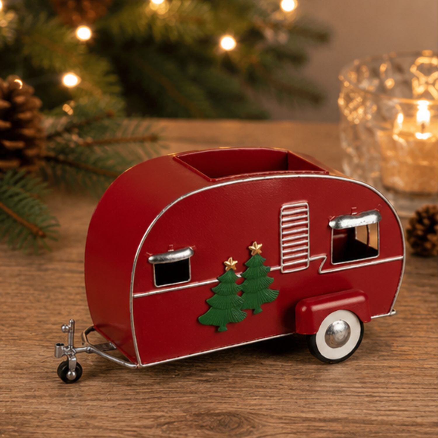 Roter Miniatur-Wohnwagen mit Tannenbaumdekor auf Holztisch, Lichter und Zweige im Hintergrund