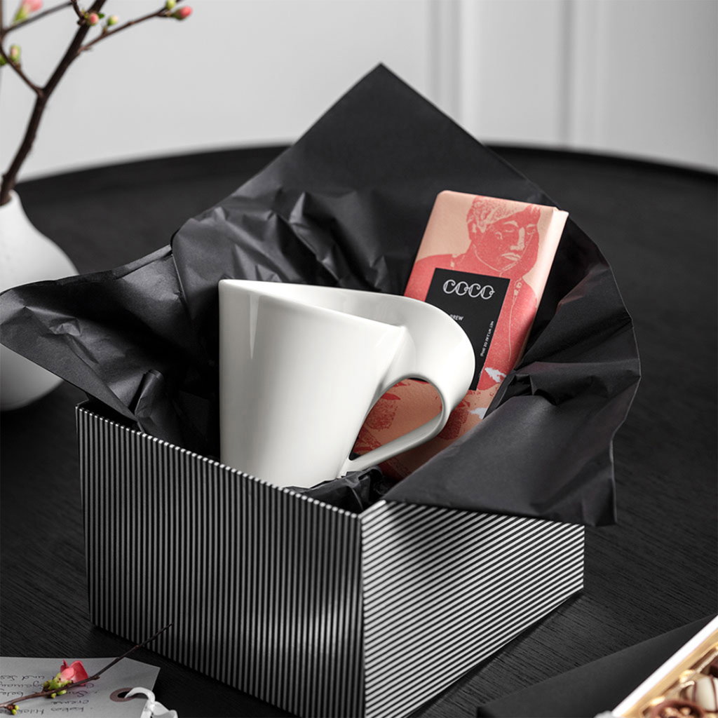 Eine weiße Tasse und eine Schokoladentafel liegen in einer schwarz-weißen Geschenkbox mit schwarzem Papier.