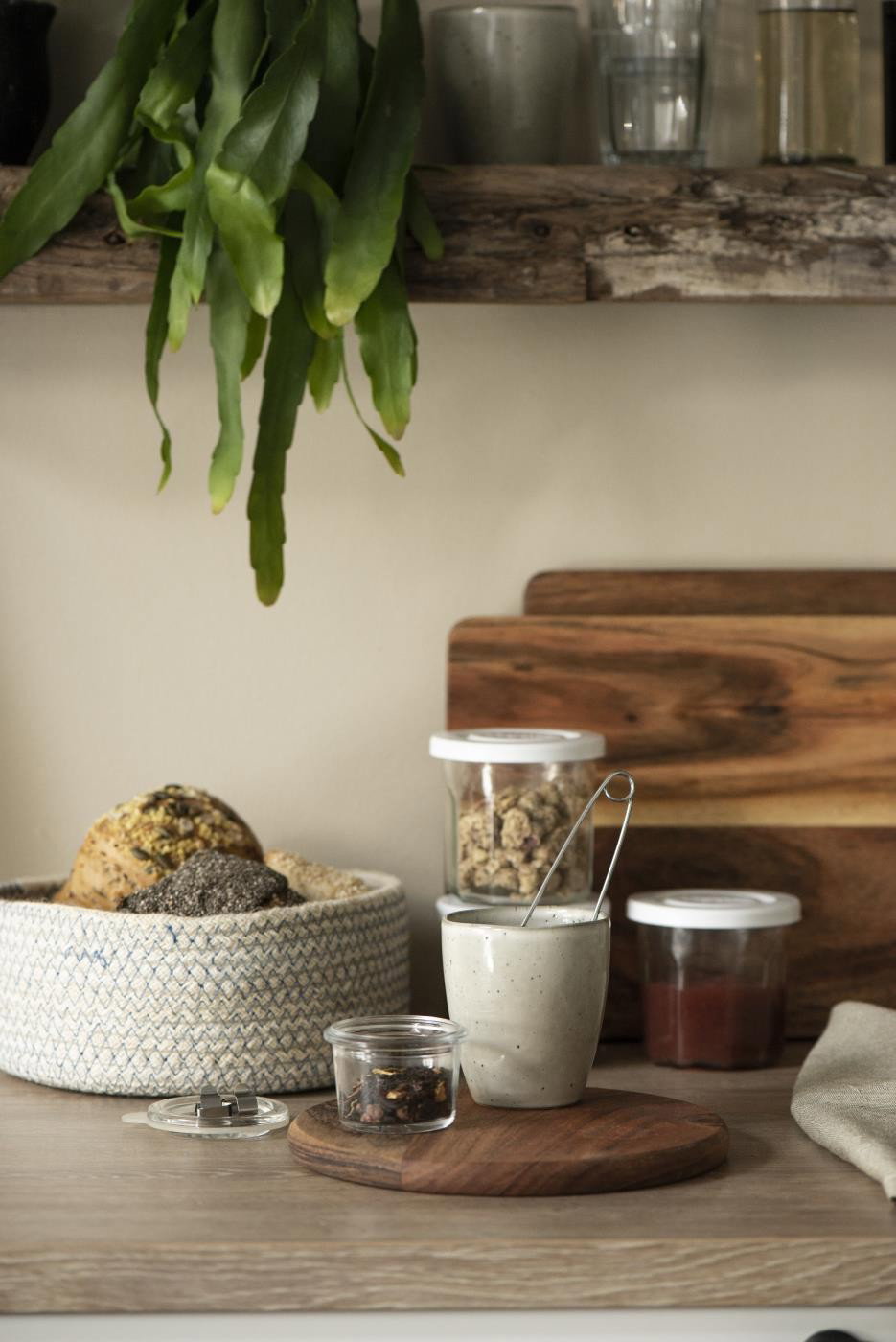 Küchenregal mit Pflanzen, Gläsern, einer Schüssel mit Brot und einer Tasse auf einem Holzbrett. Holz- und Textiloberflächen sichtbar.