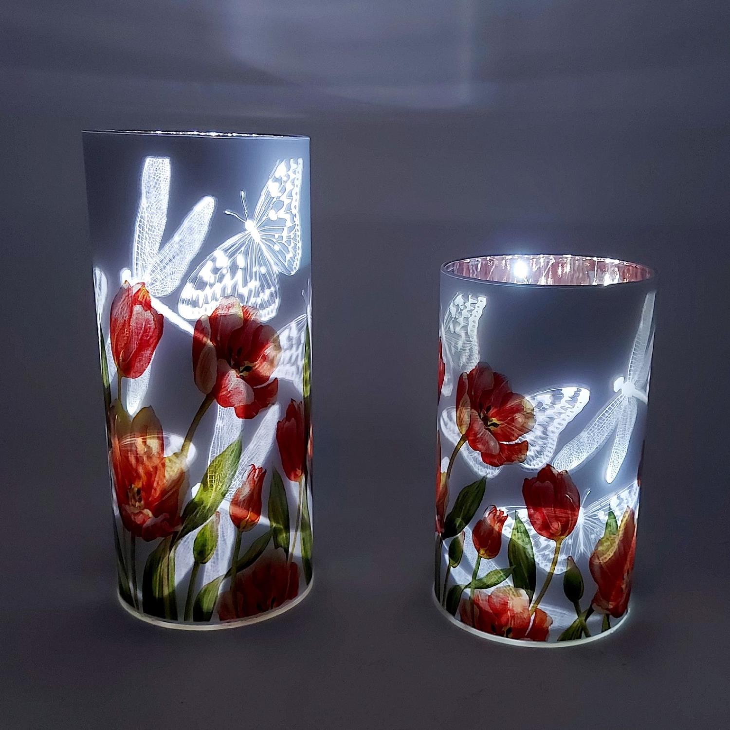 Zwei dekorative Gläser mit roten Blumenmotiven und Schmetterlingen, beleuchtet von innen, vor einem