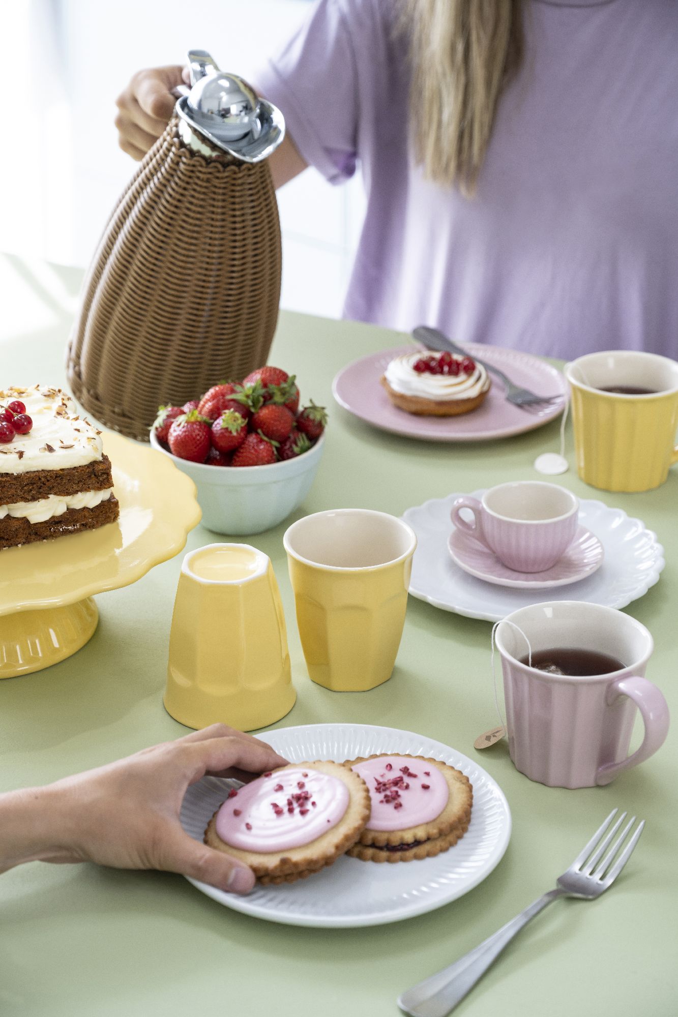 Ein gedeckter Tisch mit Kuchen, Erdbeeren, Tassen und Keksen. Eine Person gießt aus einer Kanne. Farbenfrohes Geschirr ist sichtbar.