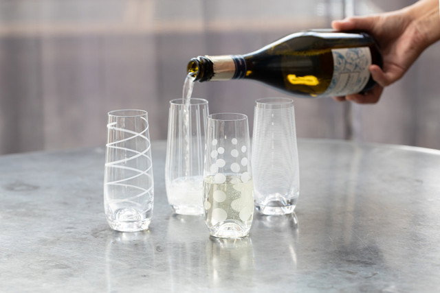 Eine Hand gießt ein Getränk aus einer Flasche in vier verschiedene Gläser auf einem Tisch.
