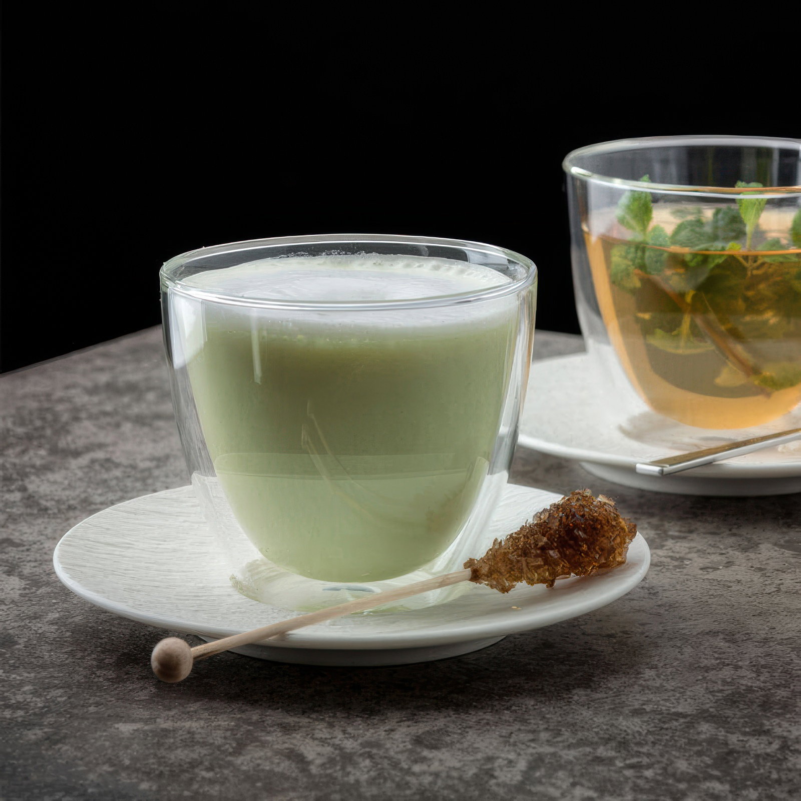 Zwei Gläser auf Untertassen: eines mit grünem Getränk und eines mit Kräutertee. Ein Zuckerstick liegt neben dem grünen Glas.