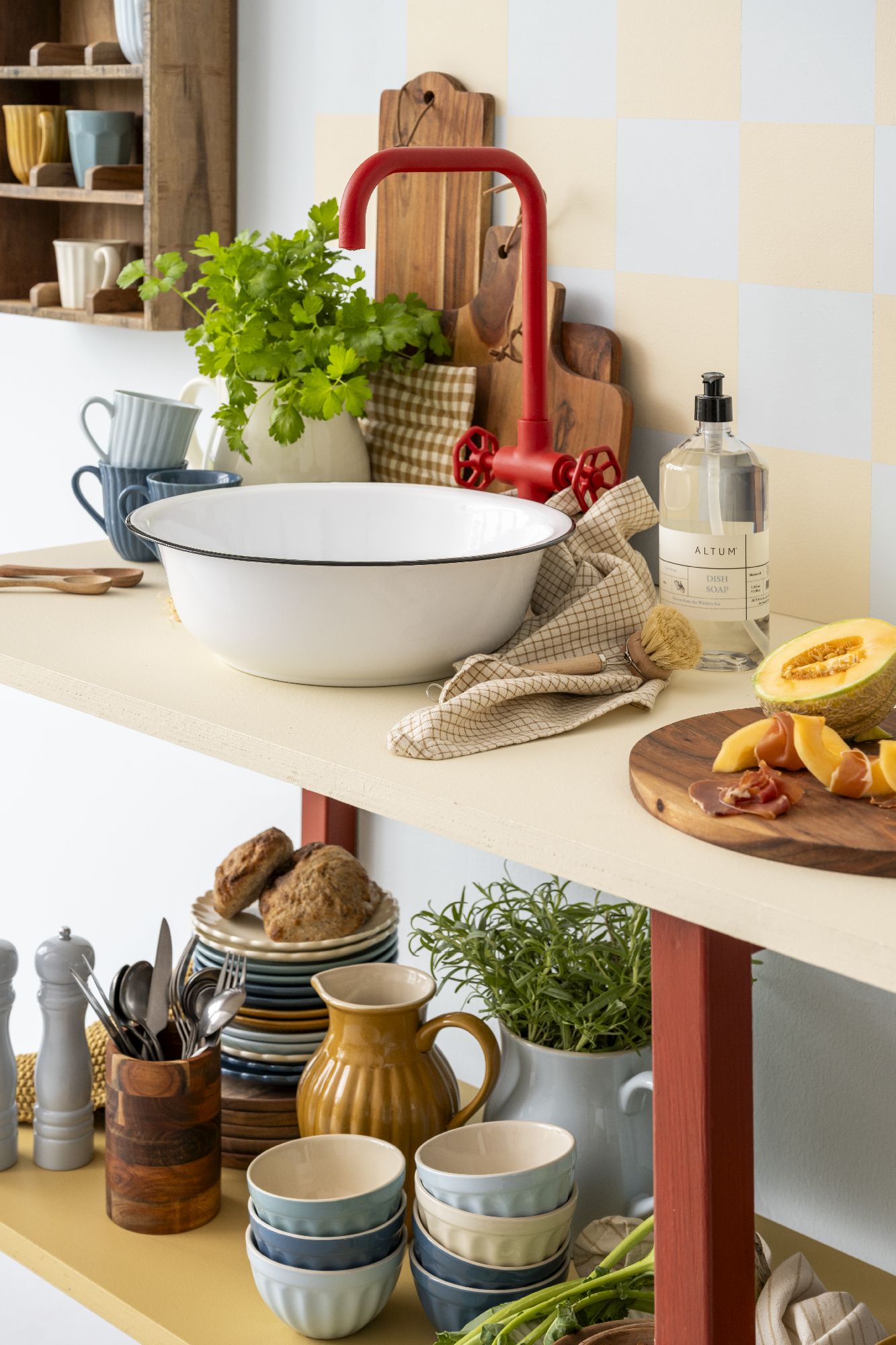 Küchenregal mit Schüssel, Kräutern, Geschirr, einem roten Wasserhahn und Lebensmitteln auf einem Holzbrett.