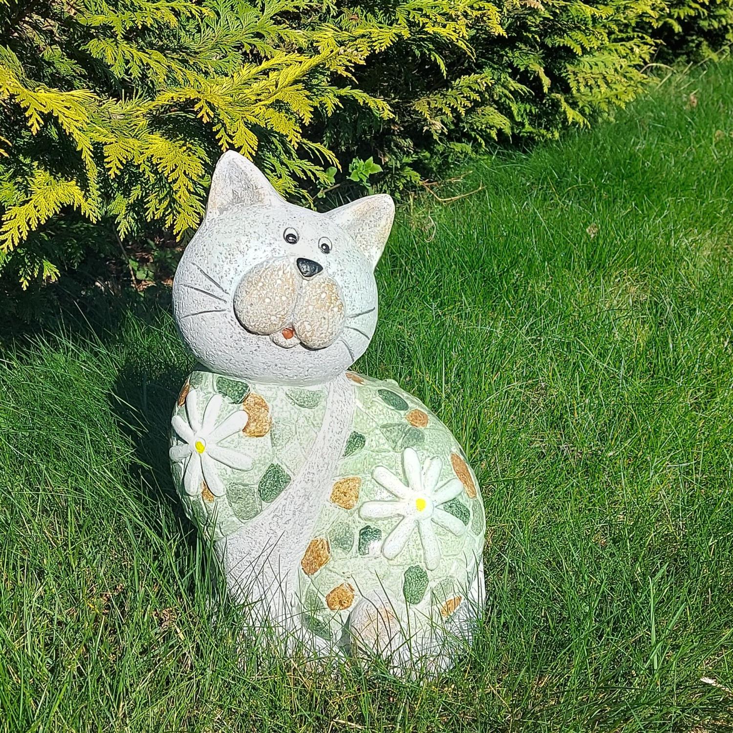 Eine dekorative Katzenfigur aus Stein mit Blumenmuster steht auf grünem Gras vor einem Nadelbaum.