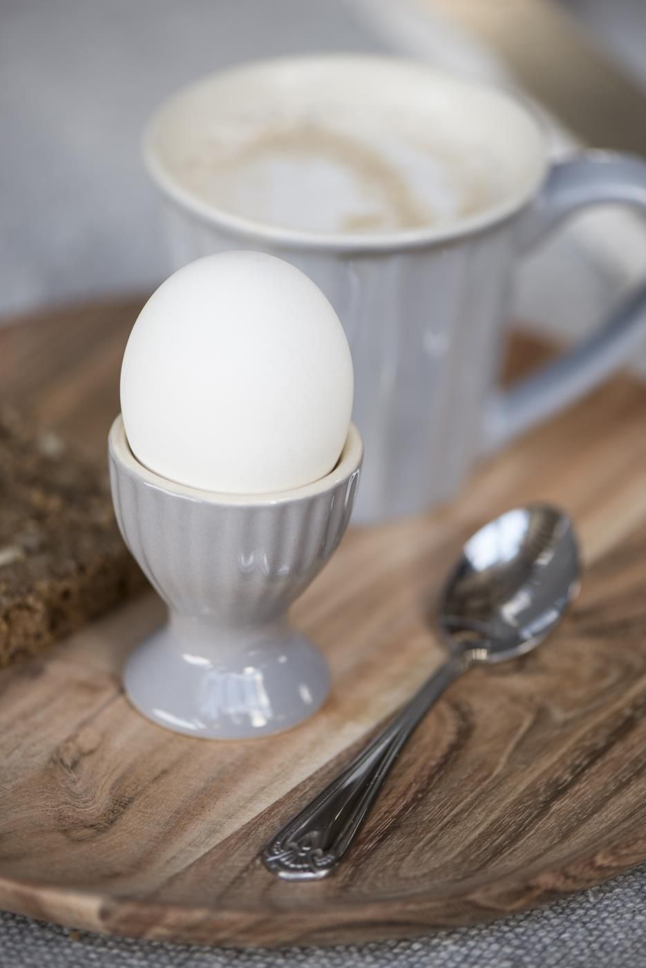 Ein gekochtes Ei in einem grauen Eierbecher, daneben ein Löffel, ein Stück Brot und eine Tasse auf einem Holzbrett.