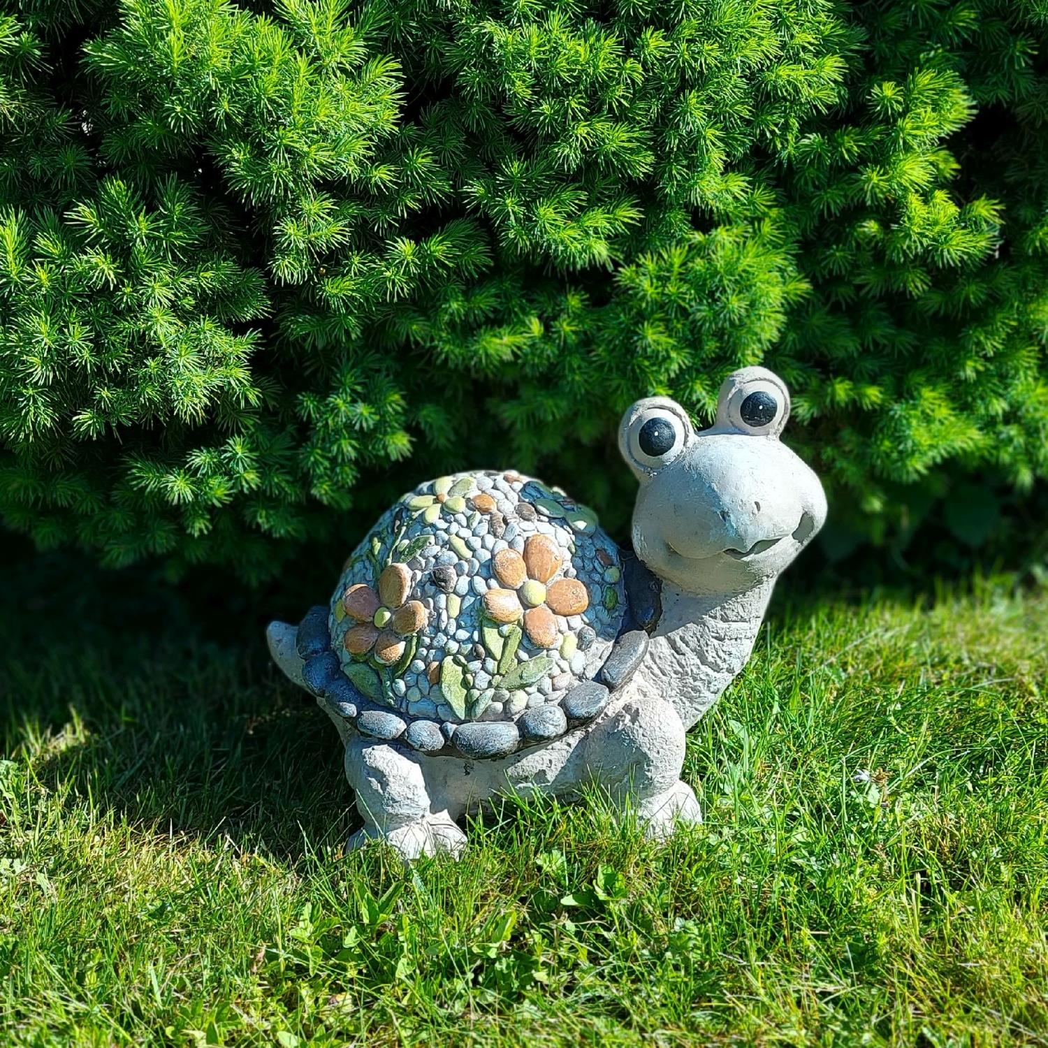 Eine bunte Steinschildkröte mit großen Augen steht auf grünem Gras vor einem dichten, grünen Busch.