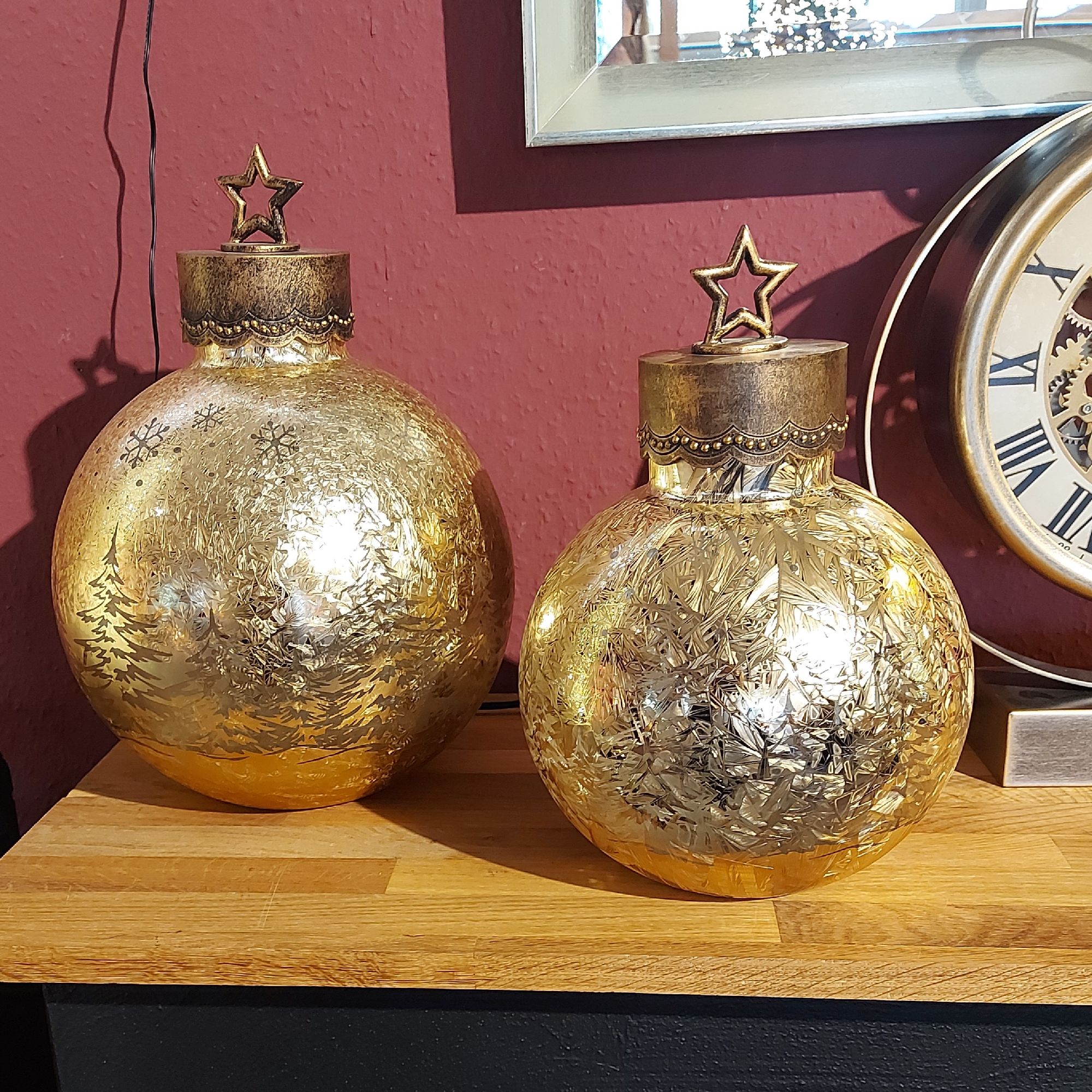 Zwei goldene Weihnachtskugeln mit Sternenaufsätzen stehen auf einem Holzregal vor einer roten Wand.