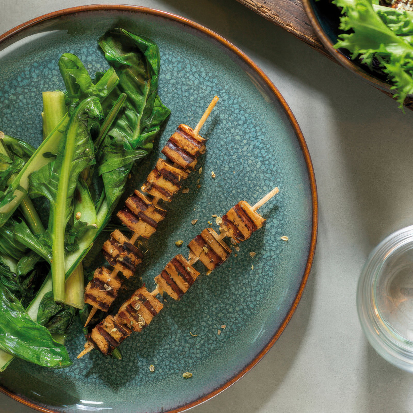 Zwei gegrillte Spieße mit mariniertem Tofu liegen auf einem blauen Teller, daneben frische grüne Blätter. Ein Glas Wasser ist sichtbar.
