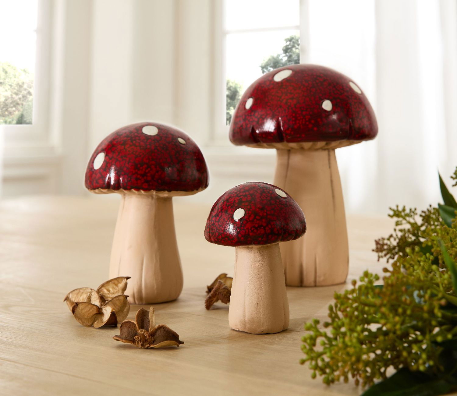 Drei dekorative Pilze mit roten Kappen und weißen Punkten stehen auf einem Tisch, umgeben von getrockneten Pflanzen.