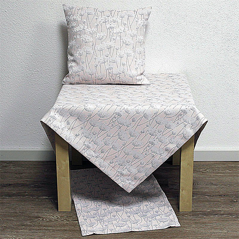 Ein Tisch mit einer Tischdecke und einem Kissen, beide mit einem floralen Muster in sanften Farben. Der Tisch steht auf einem Holzboden.