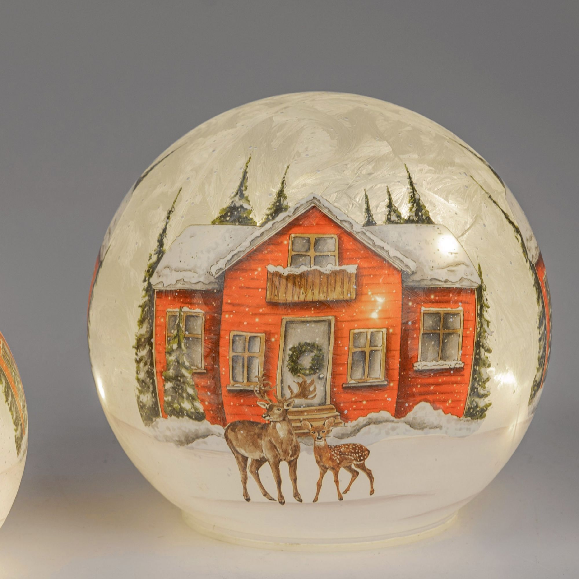 Eine Schneekugel zeigt ein rotes Haus im Schnee, umgeben von Tannenbäumen, mit zwei Rehen im Vorderg