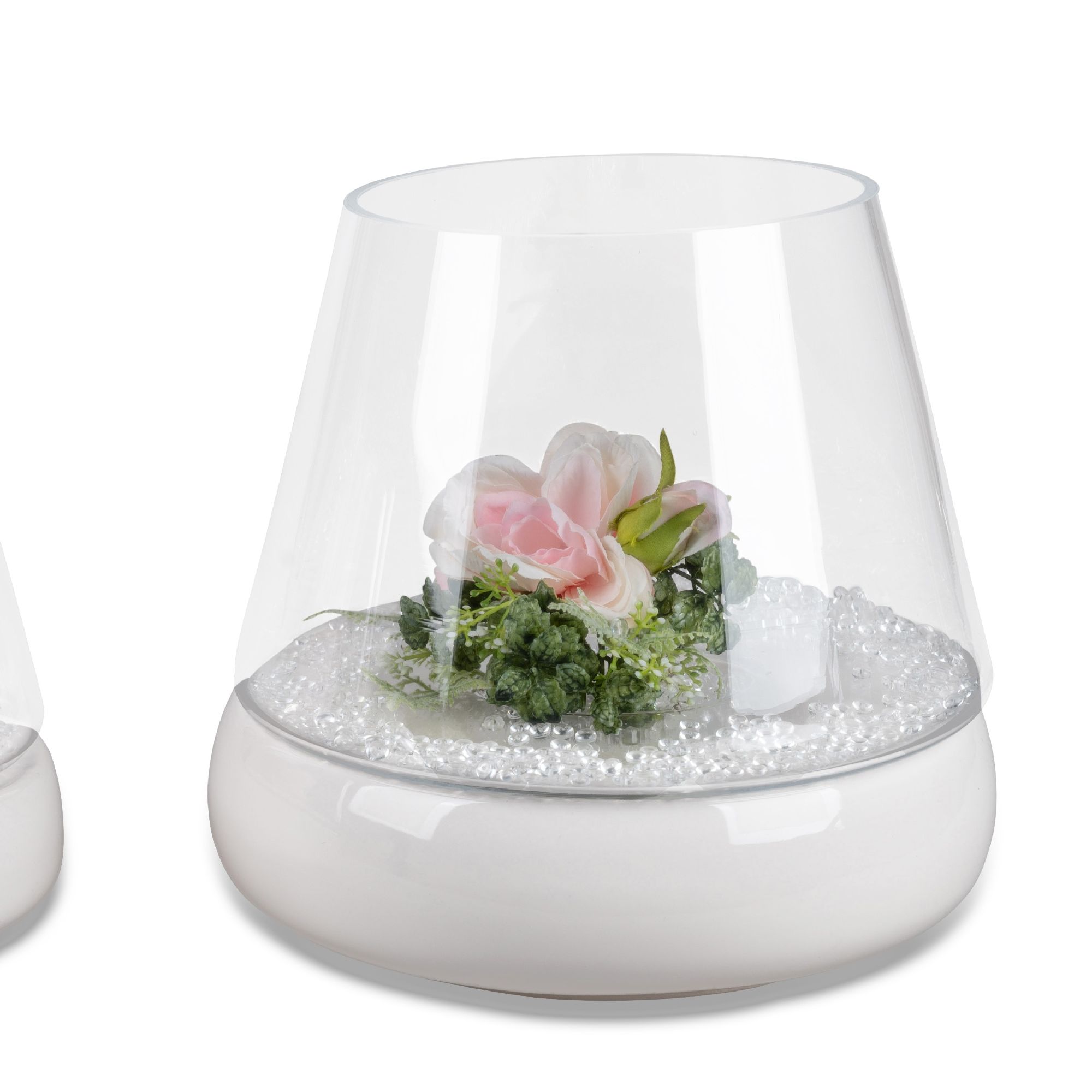 Transparente Vase mit einer rosa Blume und grünem Blattwerk, gefüllt mit weißen Perlen und auf einem weißen Sockel.