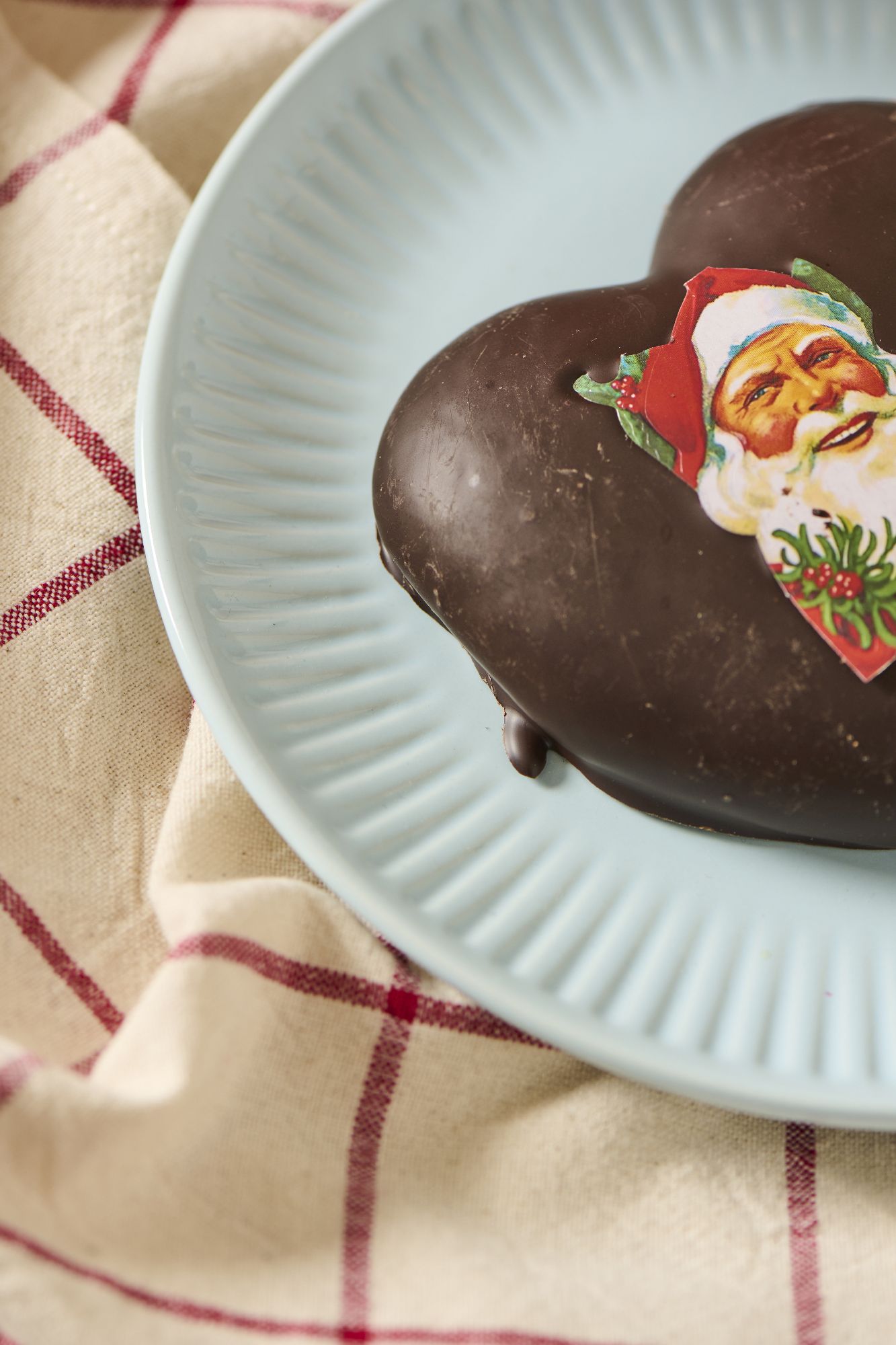 Ein herzförmiges Schokoladengebäck auf einem blauen Teller, dekoriert mit einem Weihnachtsmotiv. Im Hintergrund ein kariertes Tuch.