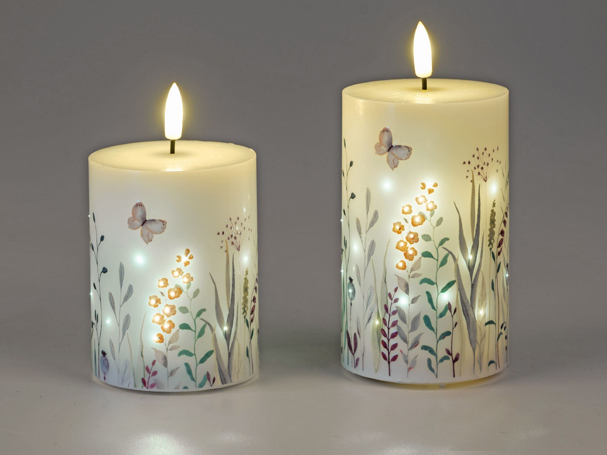 Zwei dekorative Kerzen mit floralen Motiven und Schmetterlingen, eine davon leuchtet. Hintergrund in