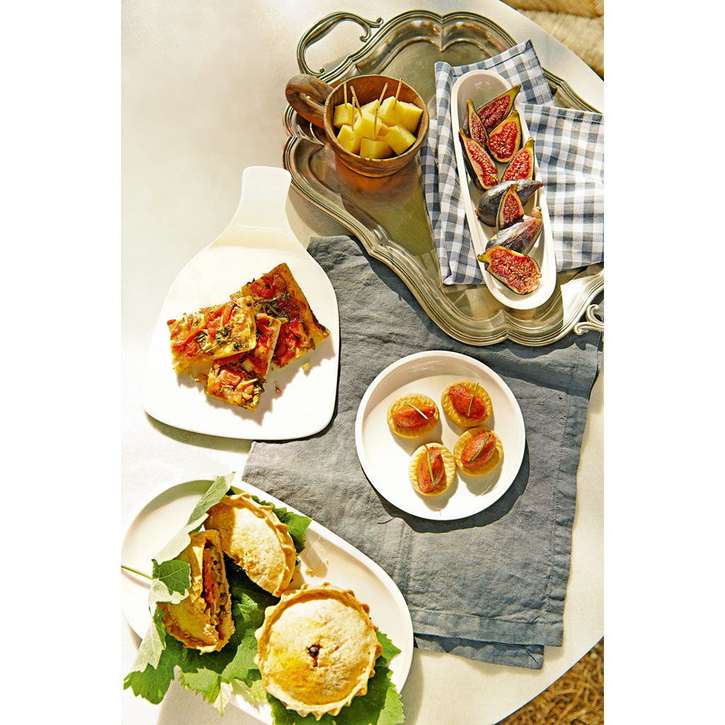 Ein Tisch mit verschiedenen Speisen: Pizza, Feigen, kleine Törtchen und Obst in einer Schale, auf einem grauen Tuch angerichtet.