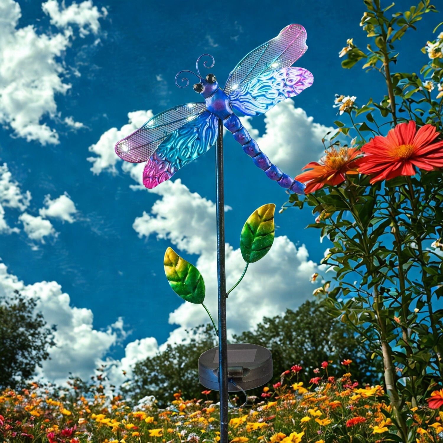 Bunte Libellen-Deko auf Stab im Blumenbeet vor blauem Himmel mit weißen Wolken