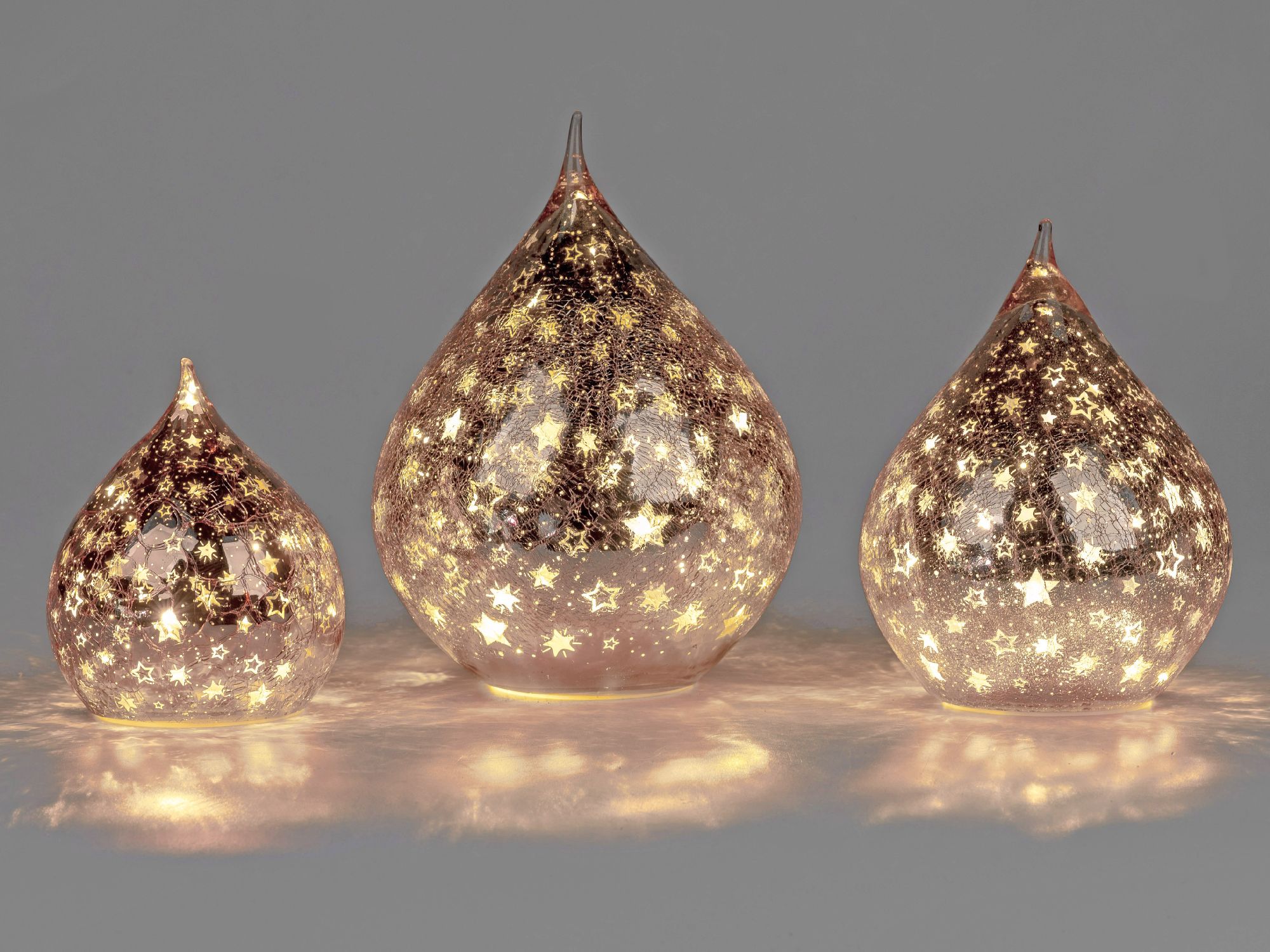 Drei gläserne Objekte in Tropfenform, beleuchtet und mit goldenen Sternenmustern verziert, stehen au