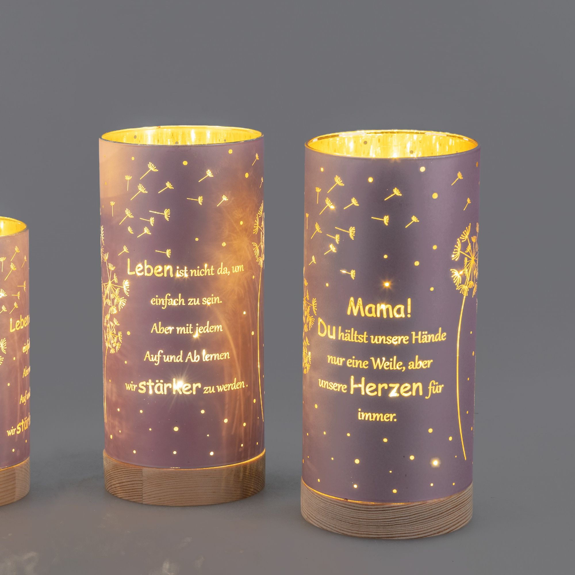 Drei dekorative Kerzen mit Sprüchen und Pusteblumen-Motiven auf lila Hintergrund, beleuchtet von innen.