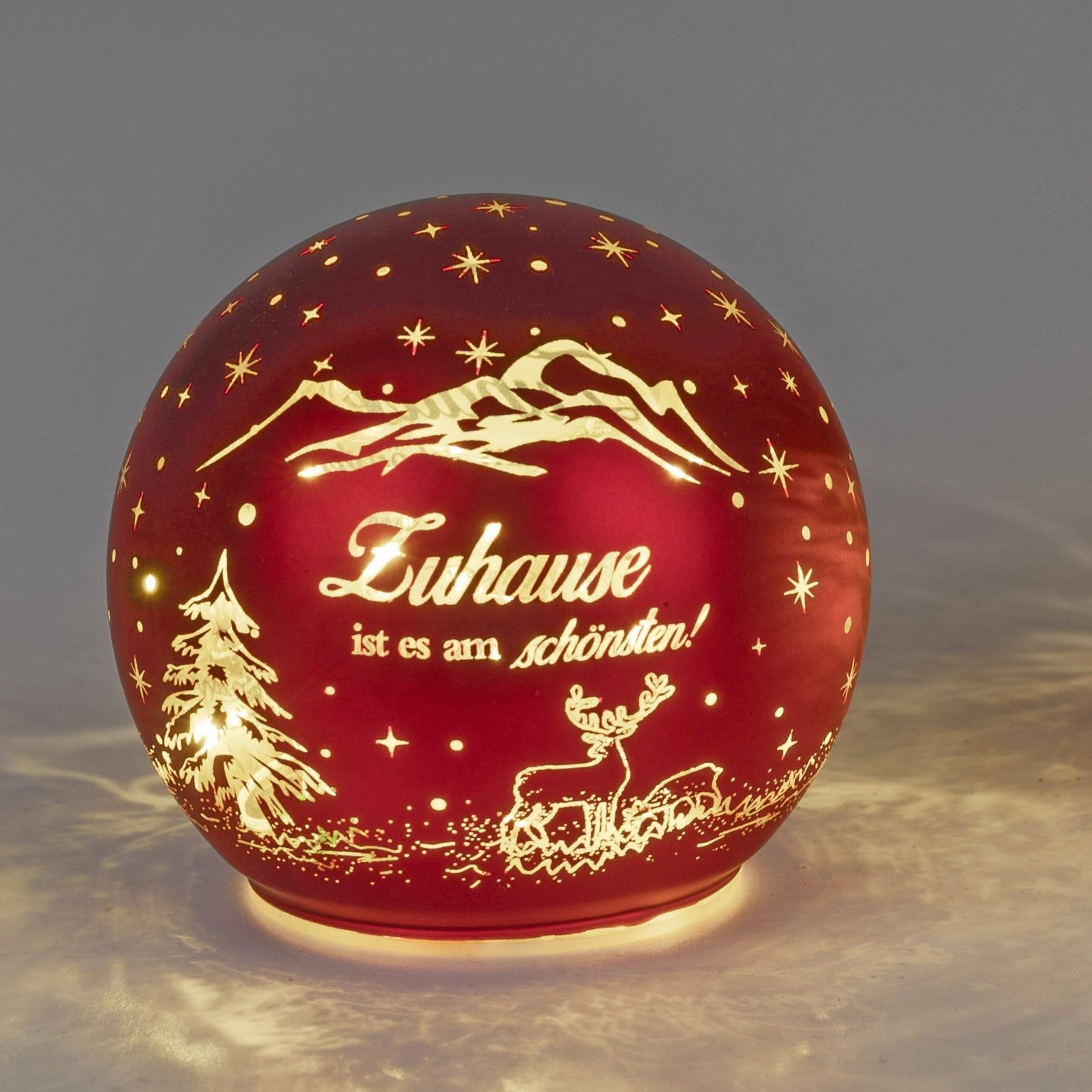 Rote Kugel mit Weihnachtsmotiven: Berge, Tannenbaum, Rentier und dem Schriftzug „Zuhause ist es am s