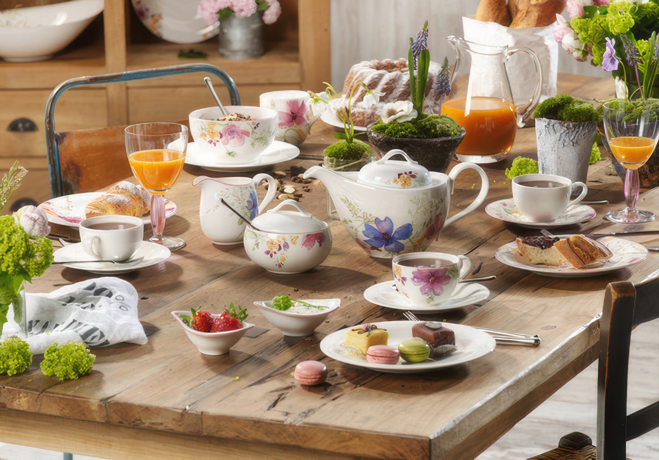 Tisch gedeckt mit Teekannen, Tassen, Gebäck, Obst und Blumen. Verschiedene Getränke in Gläsern. Holzoberfläche sichtbar.