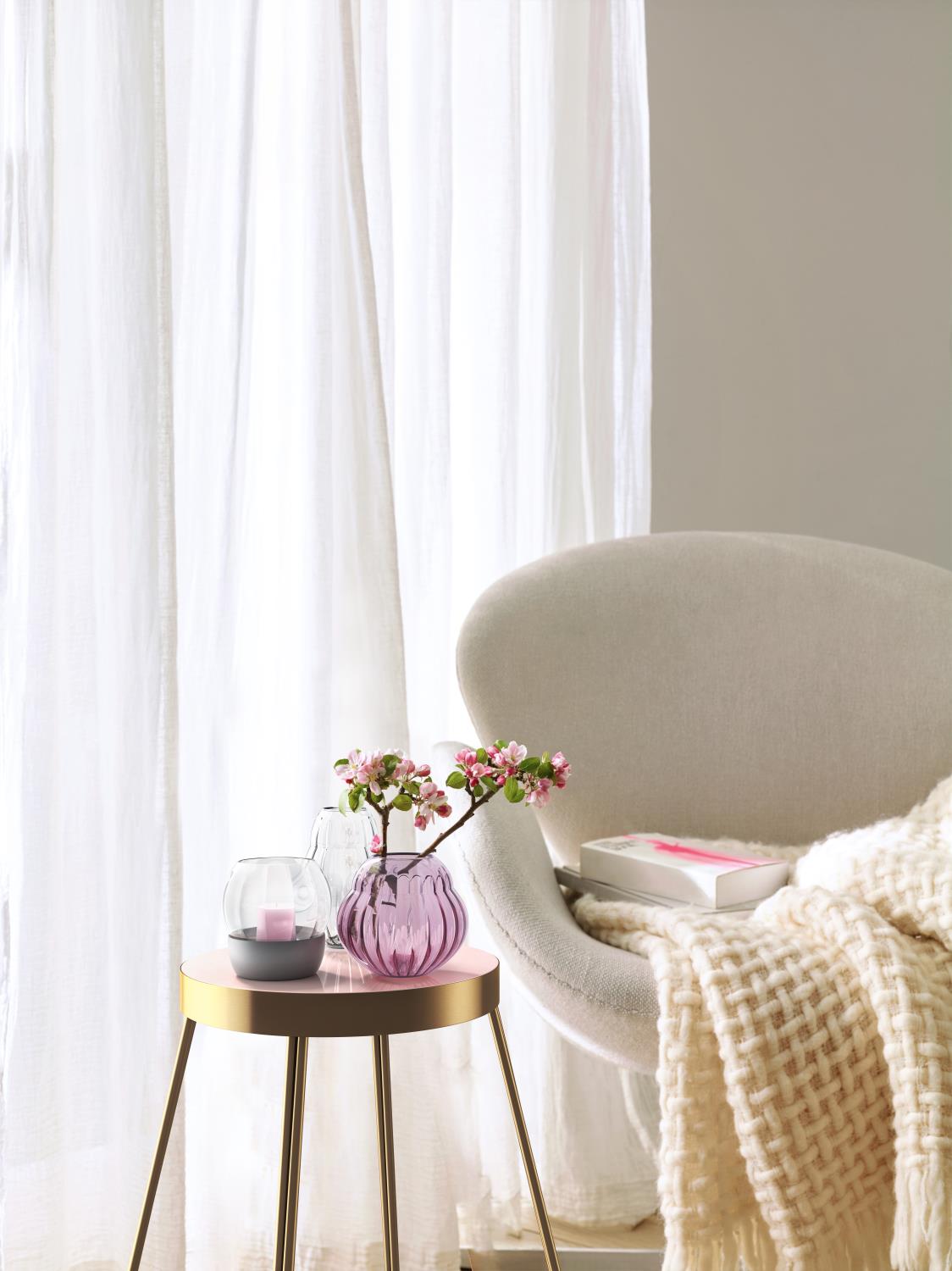 Ein beiger Sessel steht neben einem kleinen Tisch. Auf dem Tisch sind zwei Vasen mit Blumen und eine Kerze. Im Hintergrund sind Vorhänge.