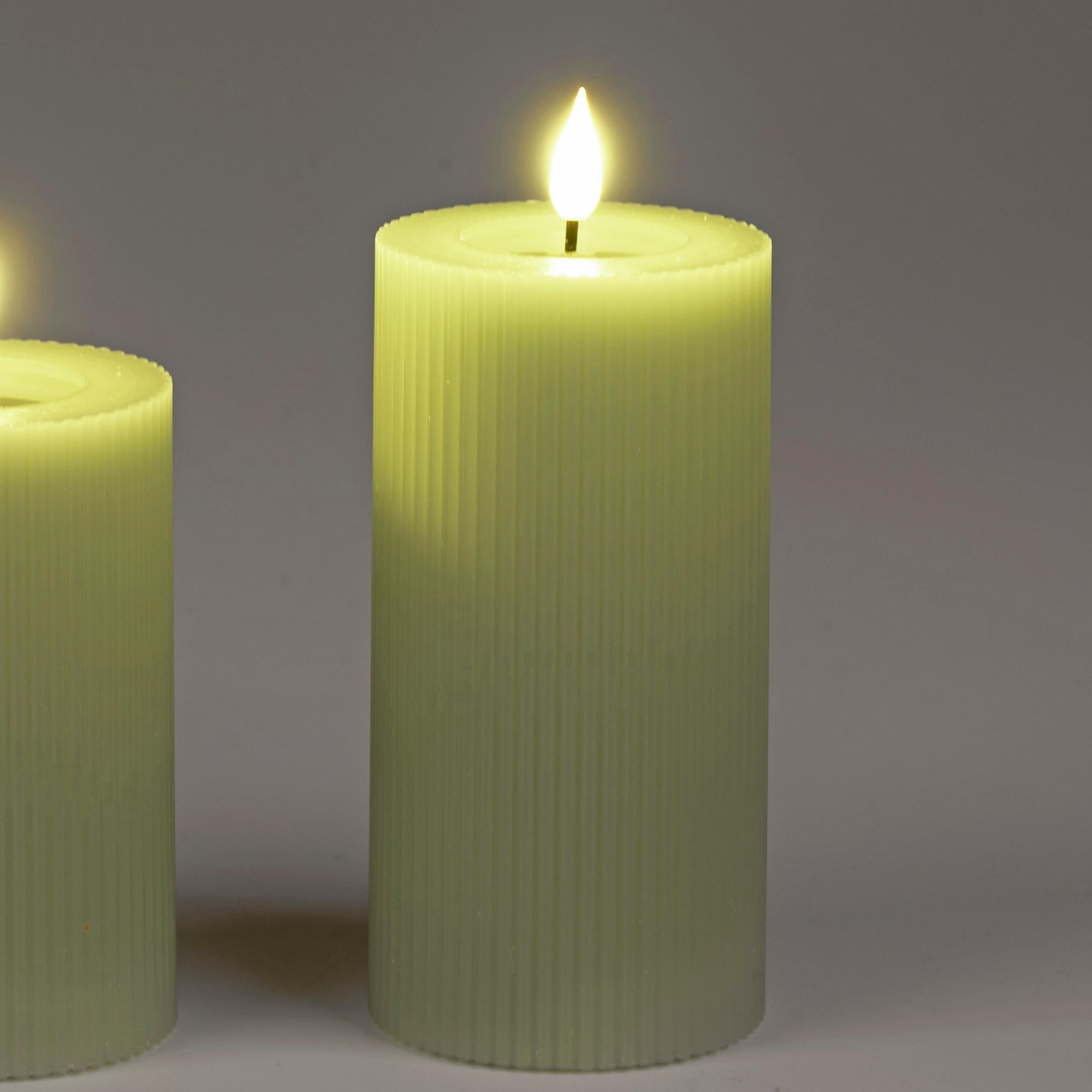 Zwei grüne, gerippte Kerzen mit flackerndem Licht auf der Oberseite, vor einem grauen Hintergrund.