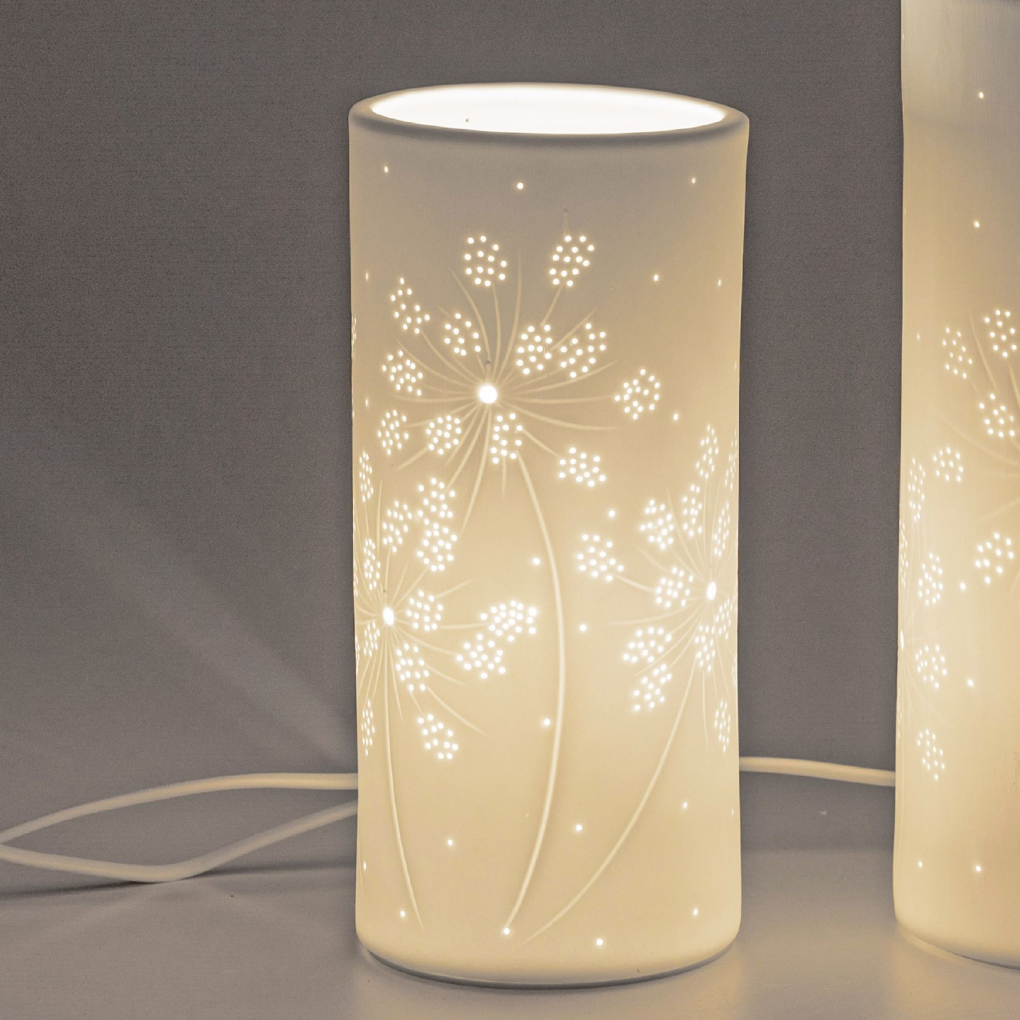 Eine weiße Tischlampe mit floralem Muster, die sanft leuchtet. Sie hat eine runde Form und ein Kabel