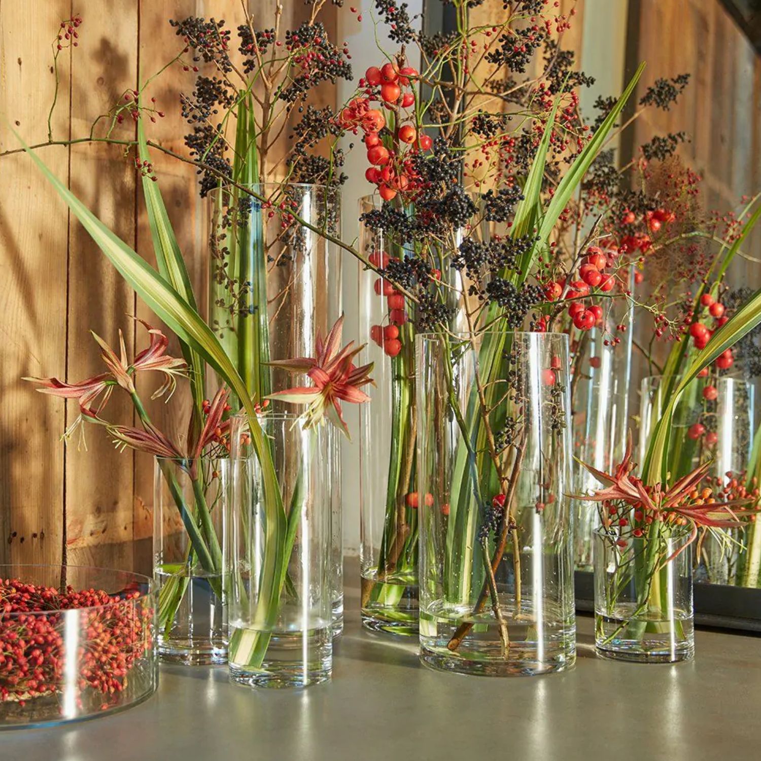 Hohe Glasvasen mit grünen Blättern, roten Beeren und zarten Blüten auf einem Tisch