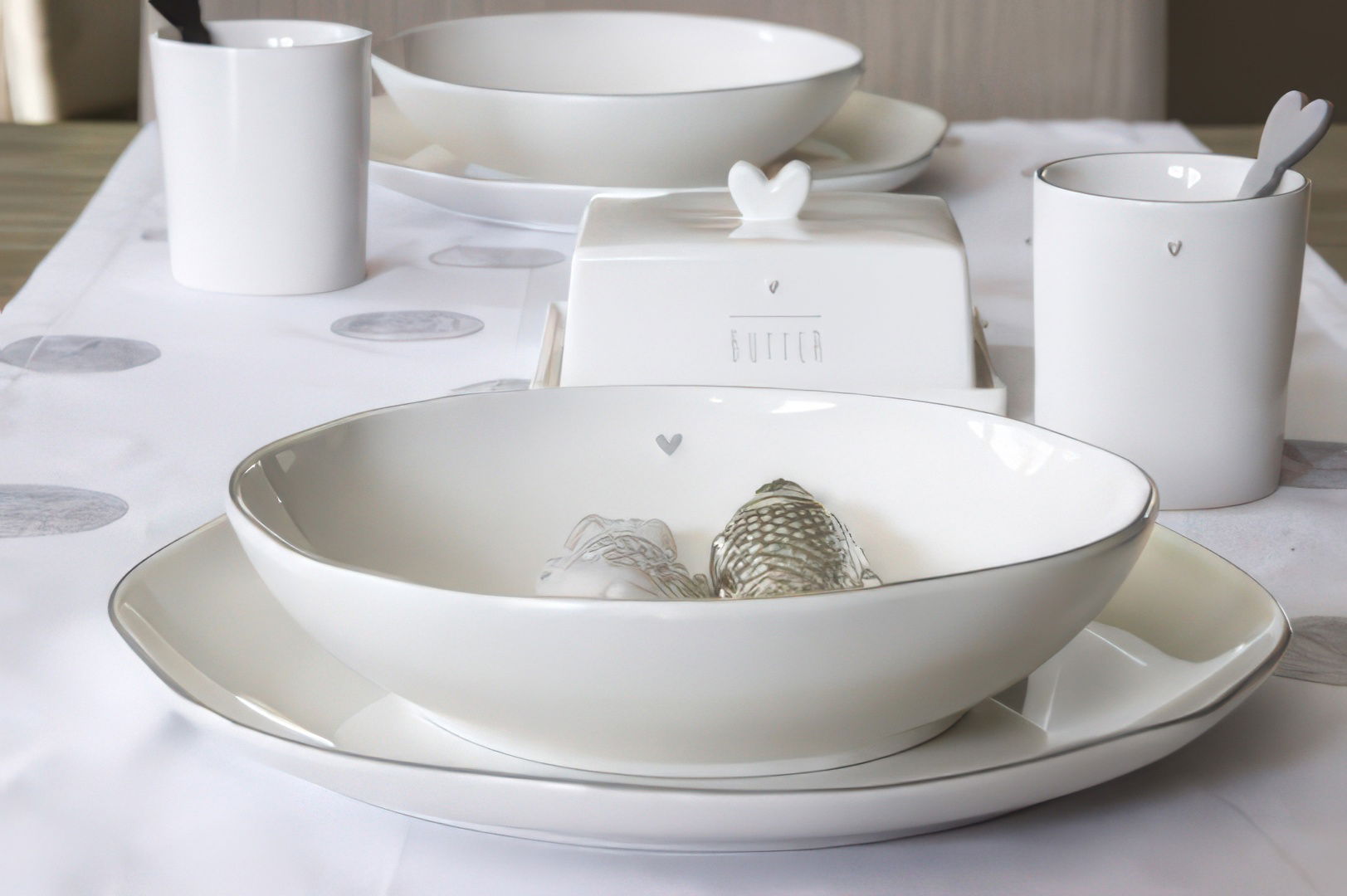 Tischgedeck mit weißen Tellern, einer Butterdose, einem Becher und einem Löffel auf einem Tisch mit einem weißen Tischläufer.