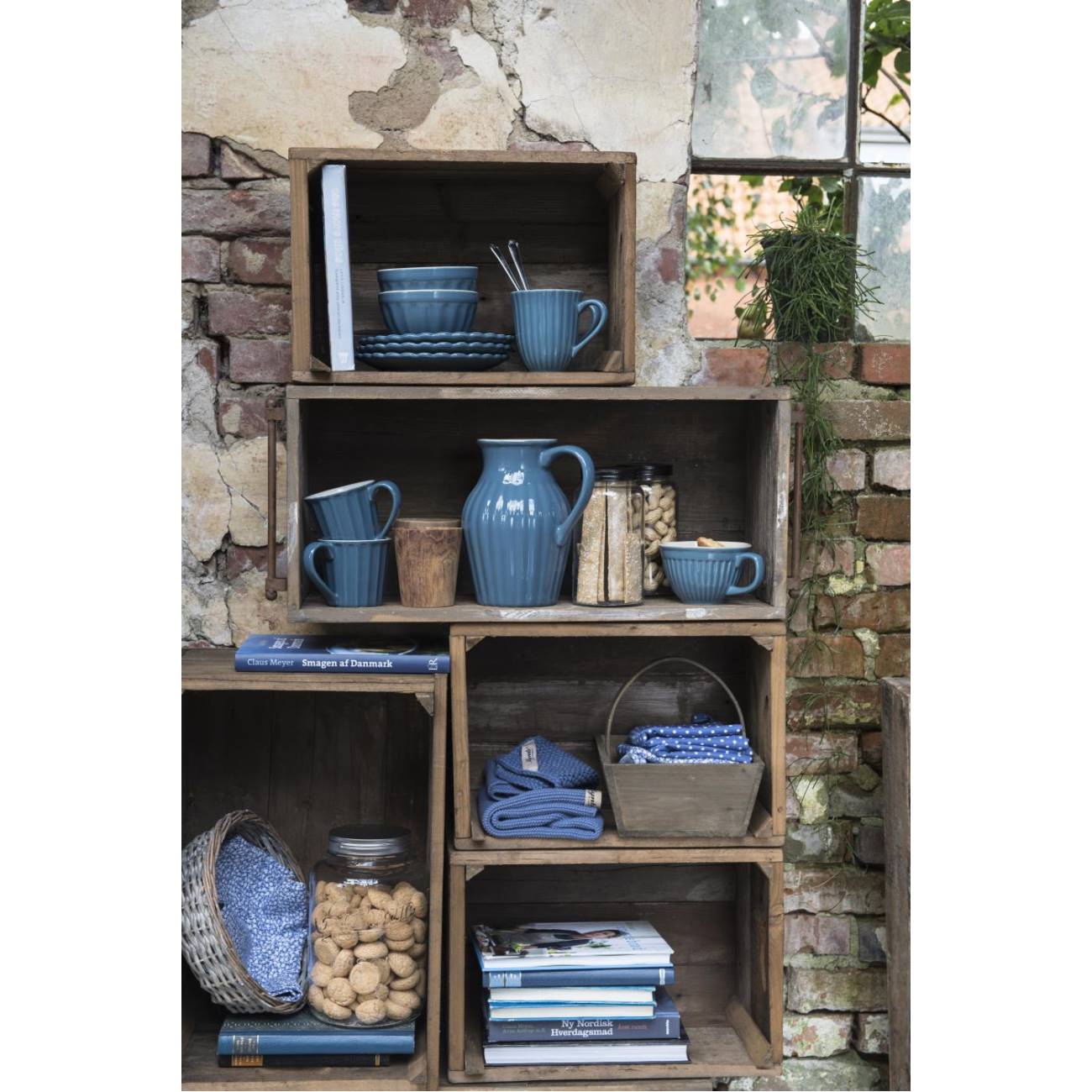 Regal aus Holz mit blauen Geschirrteilen, Gläsern, Nüssen und Büchern. Im Hintergrund eine Mauer mit Ziegelsteinen und Pflanzen.