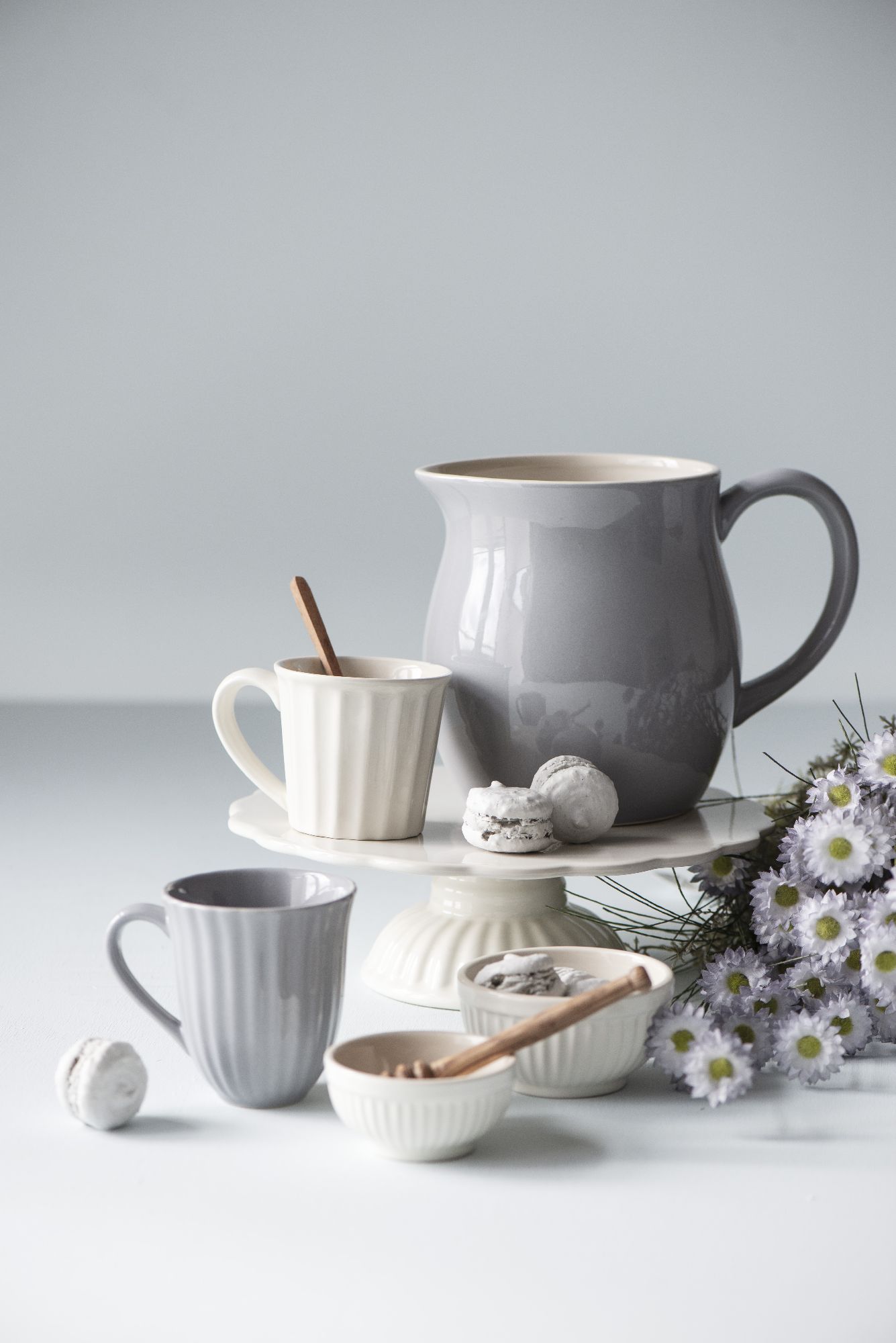 Ein grauer Krug, zwei Tassen, kleine Schalen mit Löffeln und eine Blume auf einem weißen Untergrund.