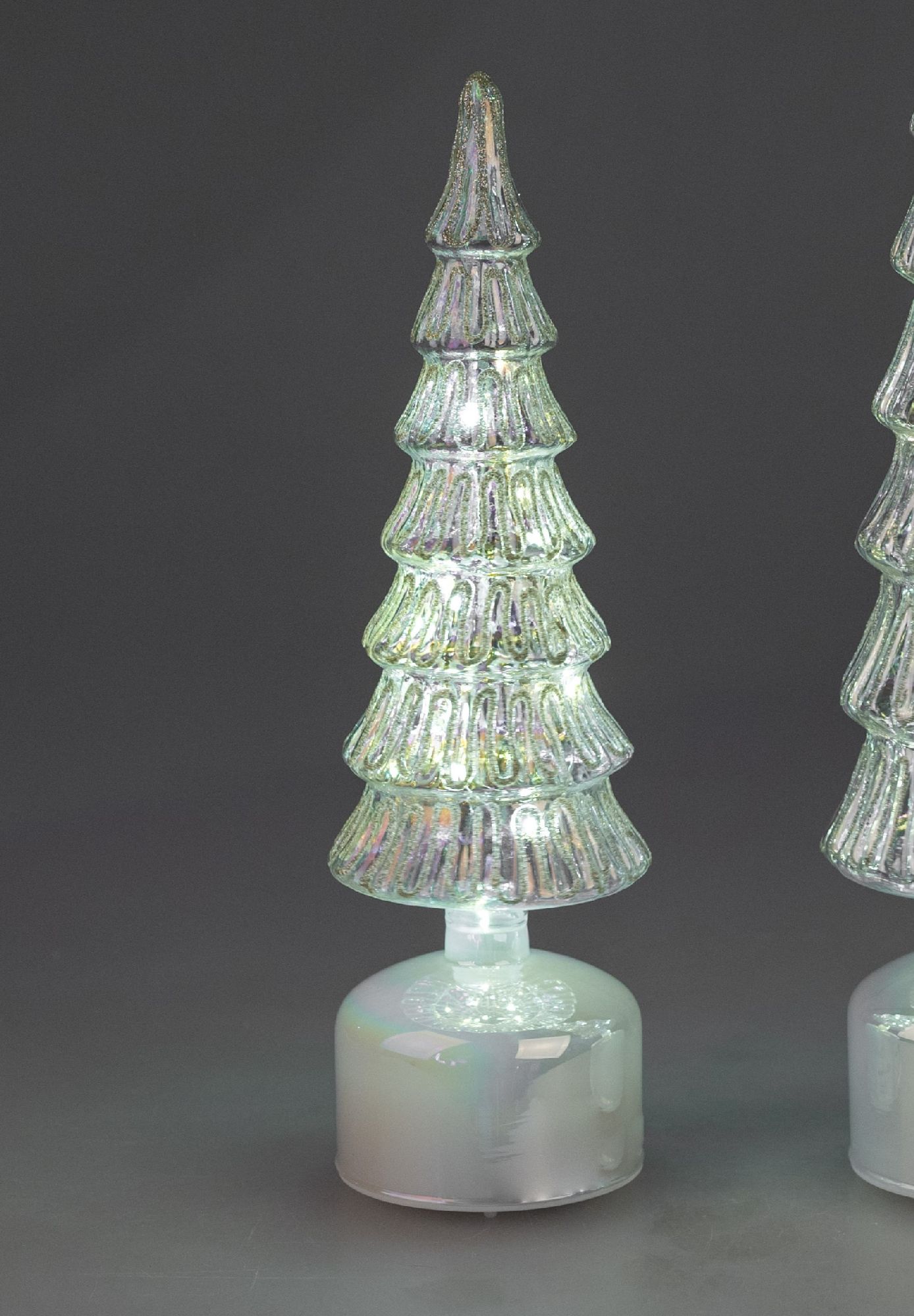 Durchsichtiger, glitzernder Weihnachtsbaum aus Glas auf einem runden, weißen Sockel, vor dunklem Hin