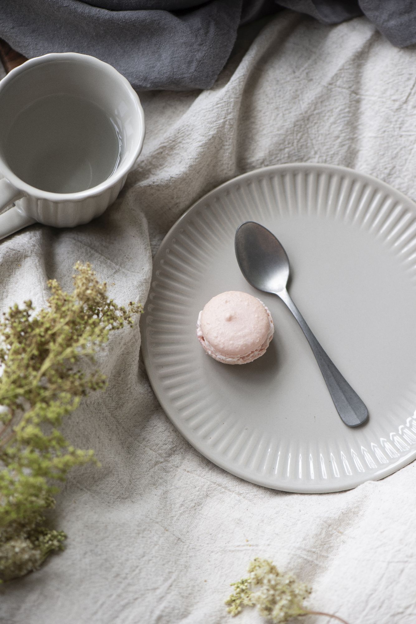 Ein grauer Teller mit einem rosa Macaron und einem Löffel. Daneben steht eine weiße Tasse mit Wasser. Im Hintergrund liegt ein graues Tuch.