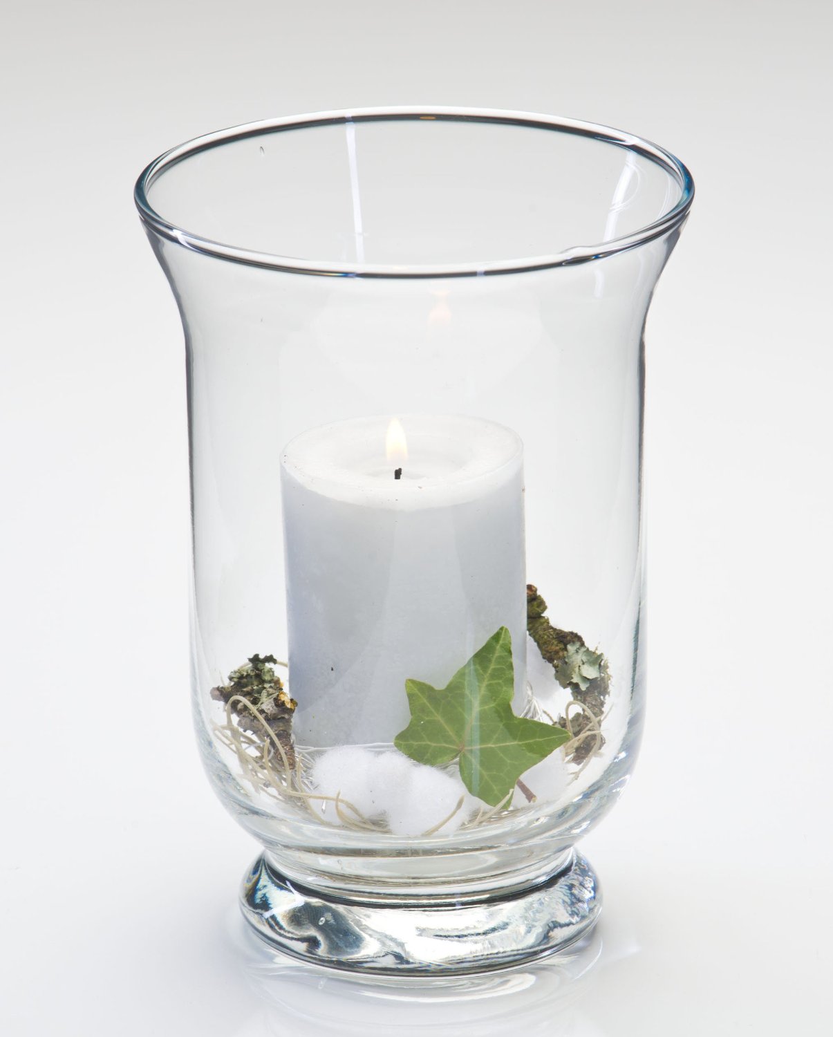 Ein transparentes Glasgefäß mit einer weißen Kerze, umgeben von Moos, Baumwolle und einem grünen Blatt.