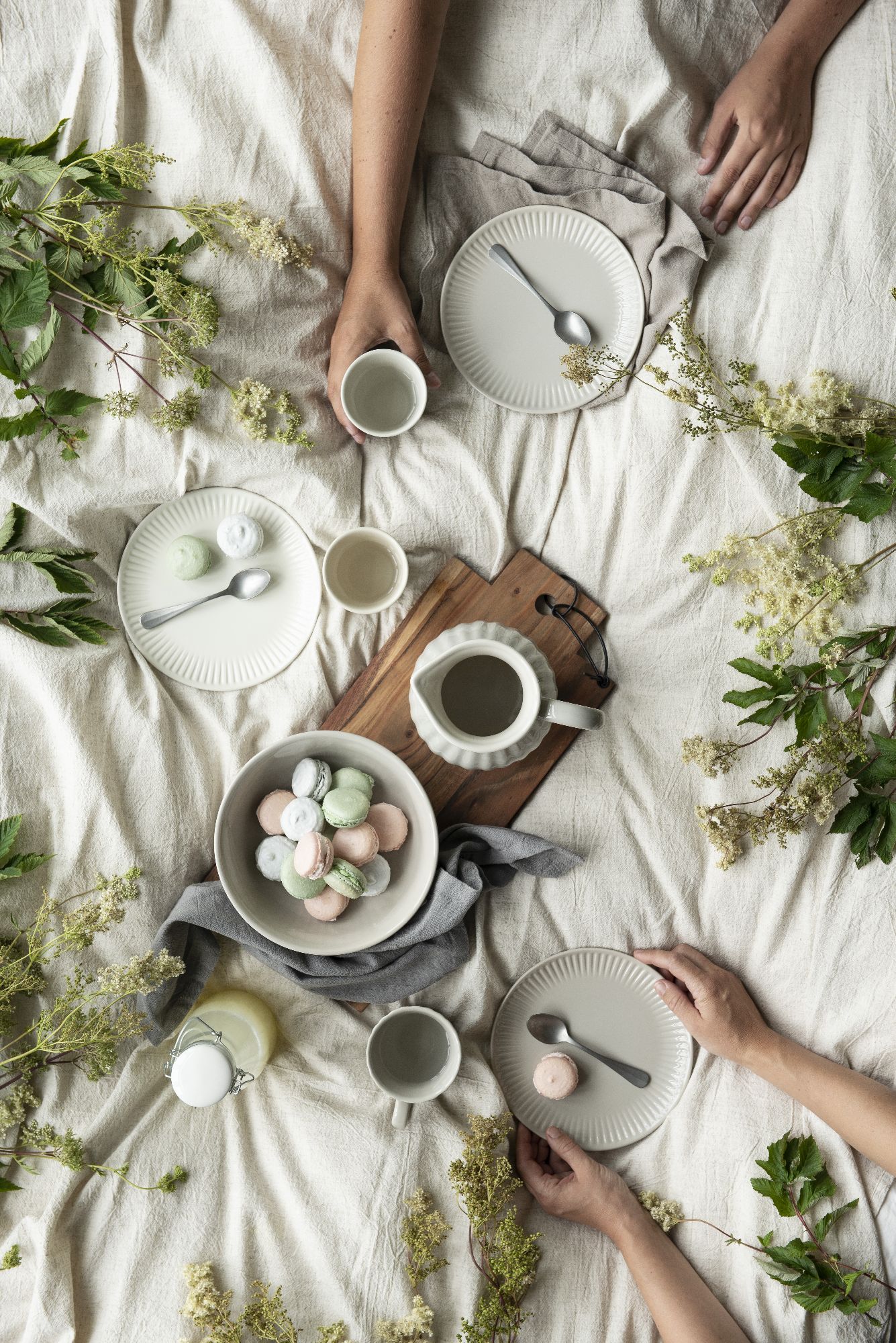 Tischgedeck mit Tellern, Tassen und einer Schüssel mit bunten Macarons, umgeben von Pflanzen und einem Tischläufer.