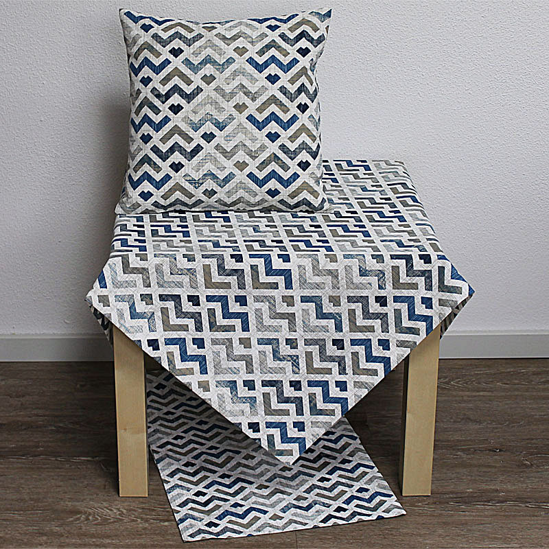 Ein Stuhl mit einem Kissen und einem Tischläufer, beide mit geometrischem Muster in Blau, Grau und Weiß. Hintergrund ist hell.