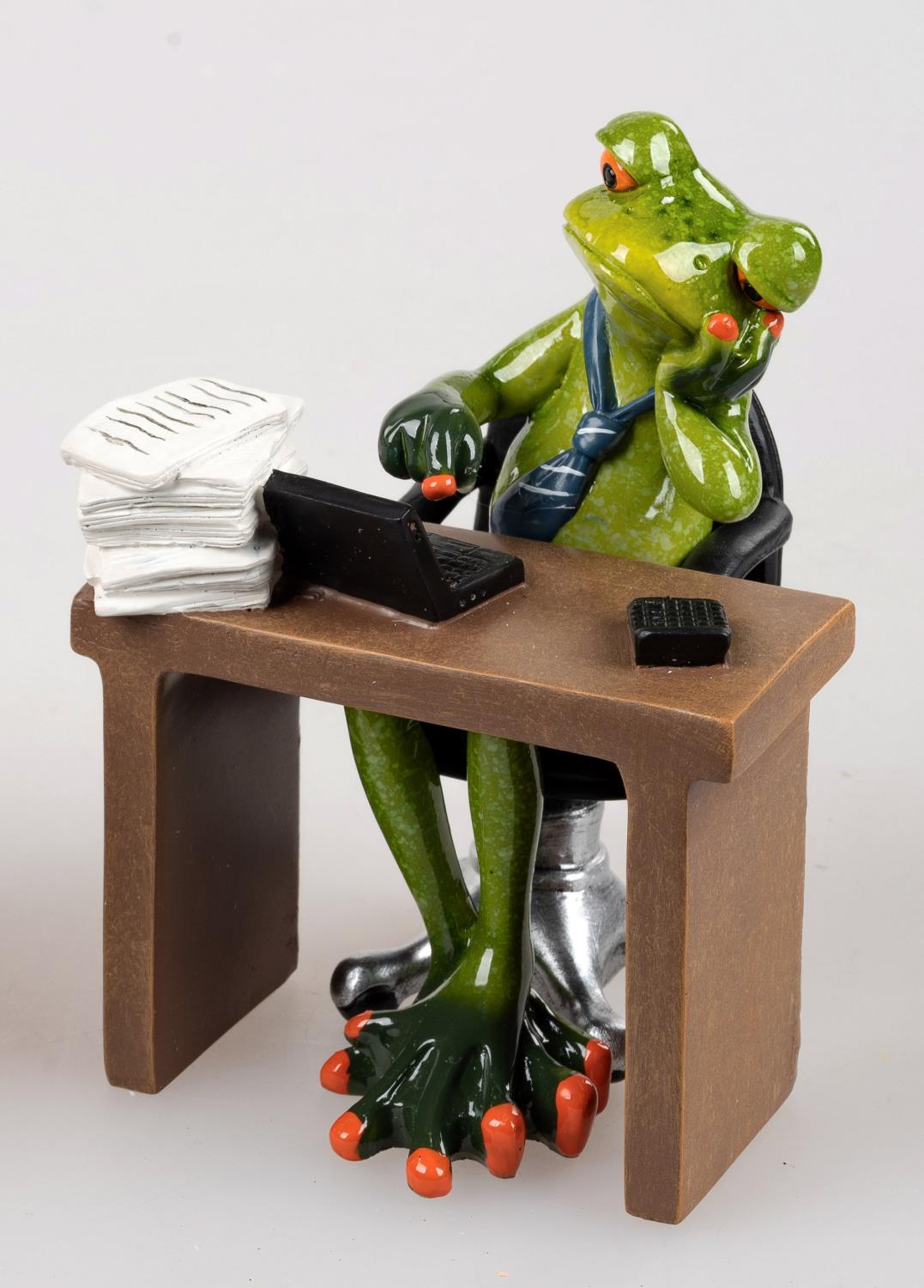 Eine grüne Froschfigur sitzt an einem Schreibtisch mit einem Laptop und Papierstapeln, trägt eine Krawatte und telefoniert.