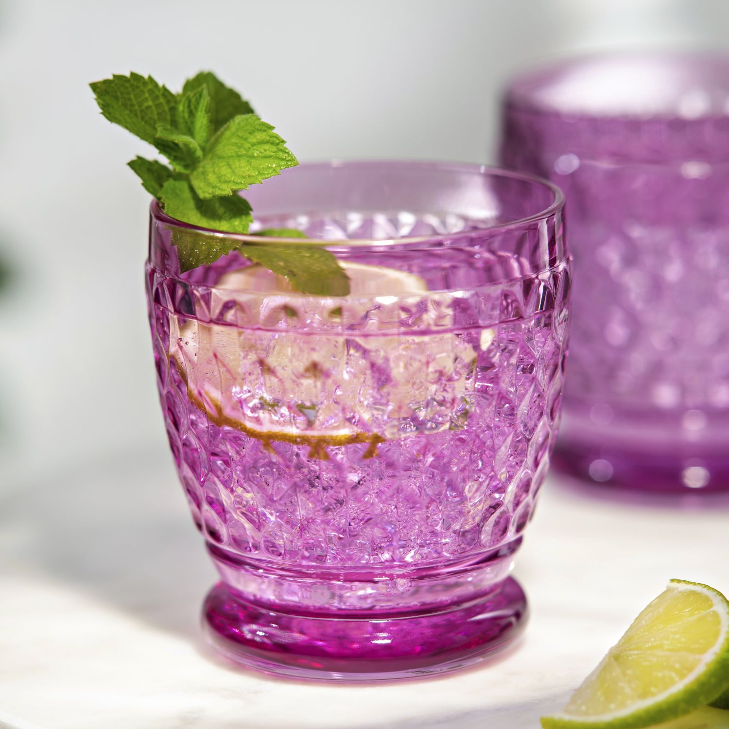 Ein lila Glas mit einem Getränk, Eiswürfeln, Limettenscheibe und Minze